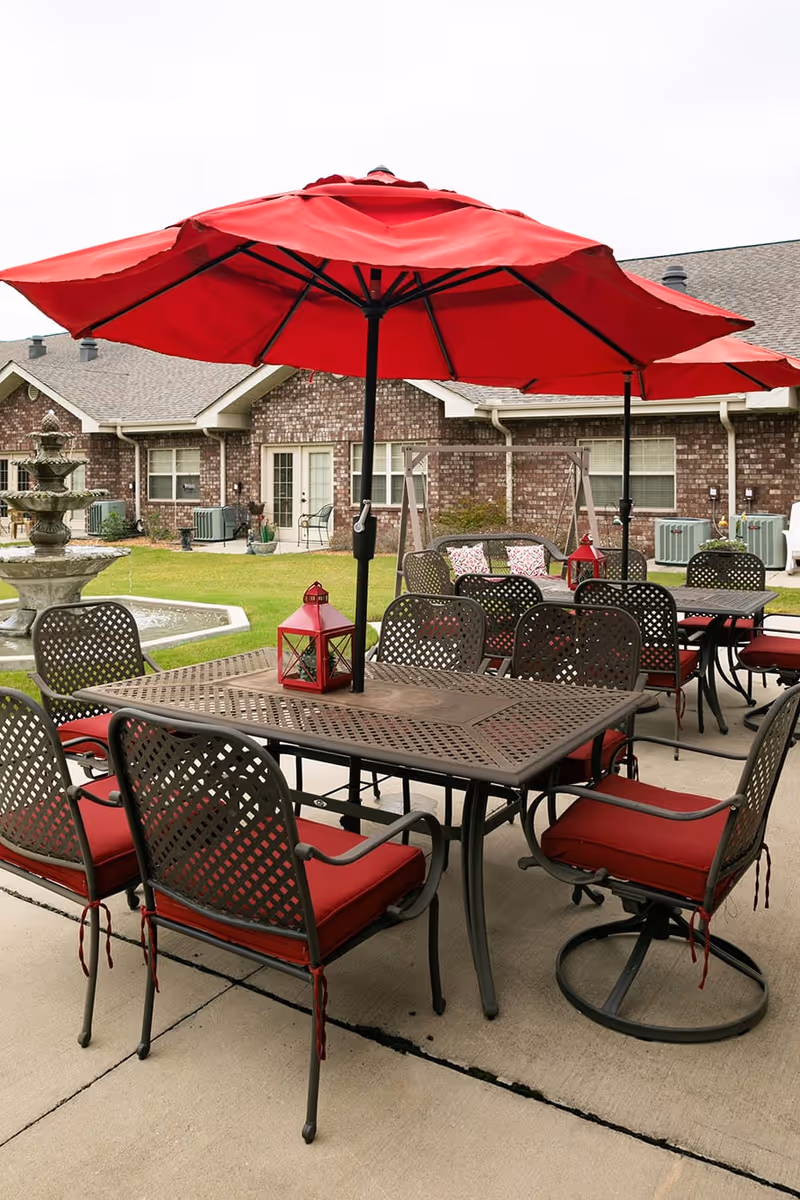 Outdoor patio area with metal tables and chairs featuring red cushions and large red umbrellas. In the background, there is a brick building with windows and doors, a swing with pillows, and a multi-tiered water fountain on a grassy lawn.