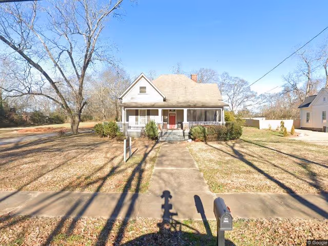 A single-story house with a front porch and a red door, situated on a grassy lot with leafless trees and a concrete walkway leading to the entrance. The sky is clear and blue.