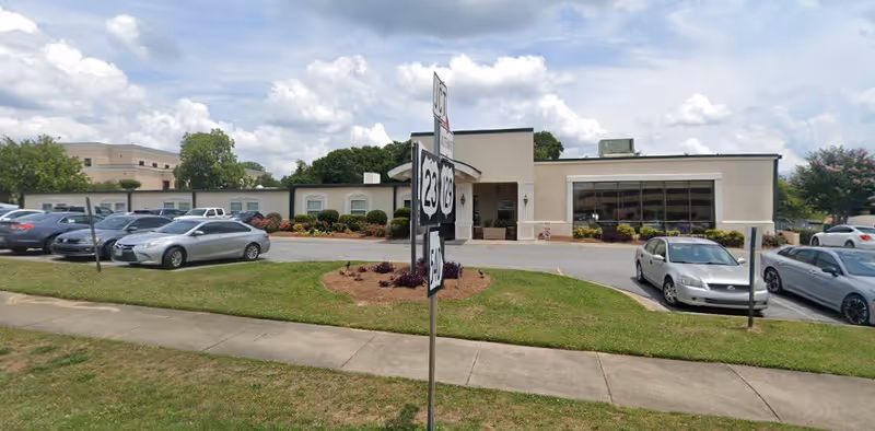 Exterior view of a single-story building with a parking lot in front, several cars parked, and a road sign showing route numbers 23 and 129. The building has a beige facade with large windows and some landscaping with bushes and flowers. The sky is partly cloudy.