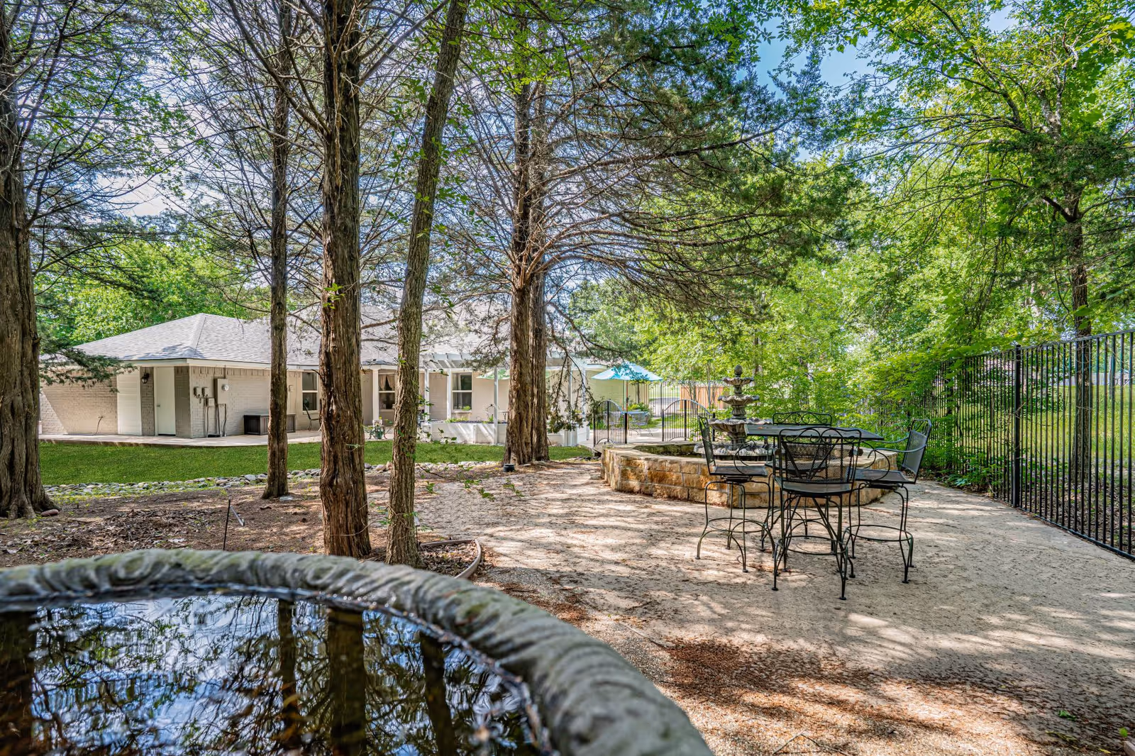 Outdoor garden area at Silver Leaf Assisted Living & Memory Care featuring tall trees, a stone water fountain, black metal patio table and chairs, and a white building in the background under a clear blue sky.