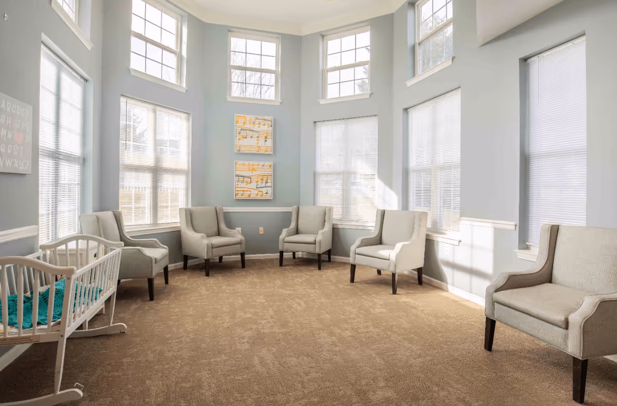 A bright and spacious sitting room with six beige armchairs arranged in a semi-circle along the walls. The room features large windows with white blinds allowing natural light to fill the space. There are two framed musical note artworks on the wall and a white wooden cradle with a teal blanket inside on the left side. The walls are painted light blue and the floor is carpeted in a light brown color.