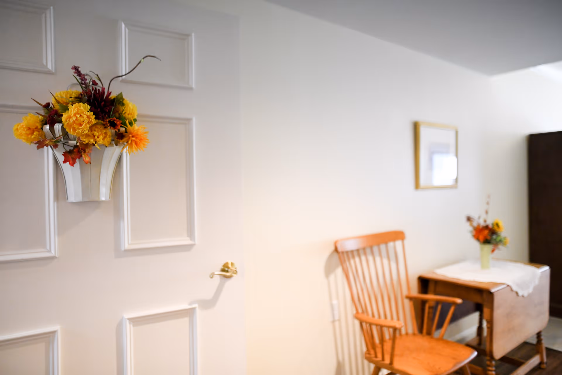 A white paneled door with a hanging floral arrangement next to a wooden chair and a small side table holding a vase of flowers in a cozy sitting area.