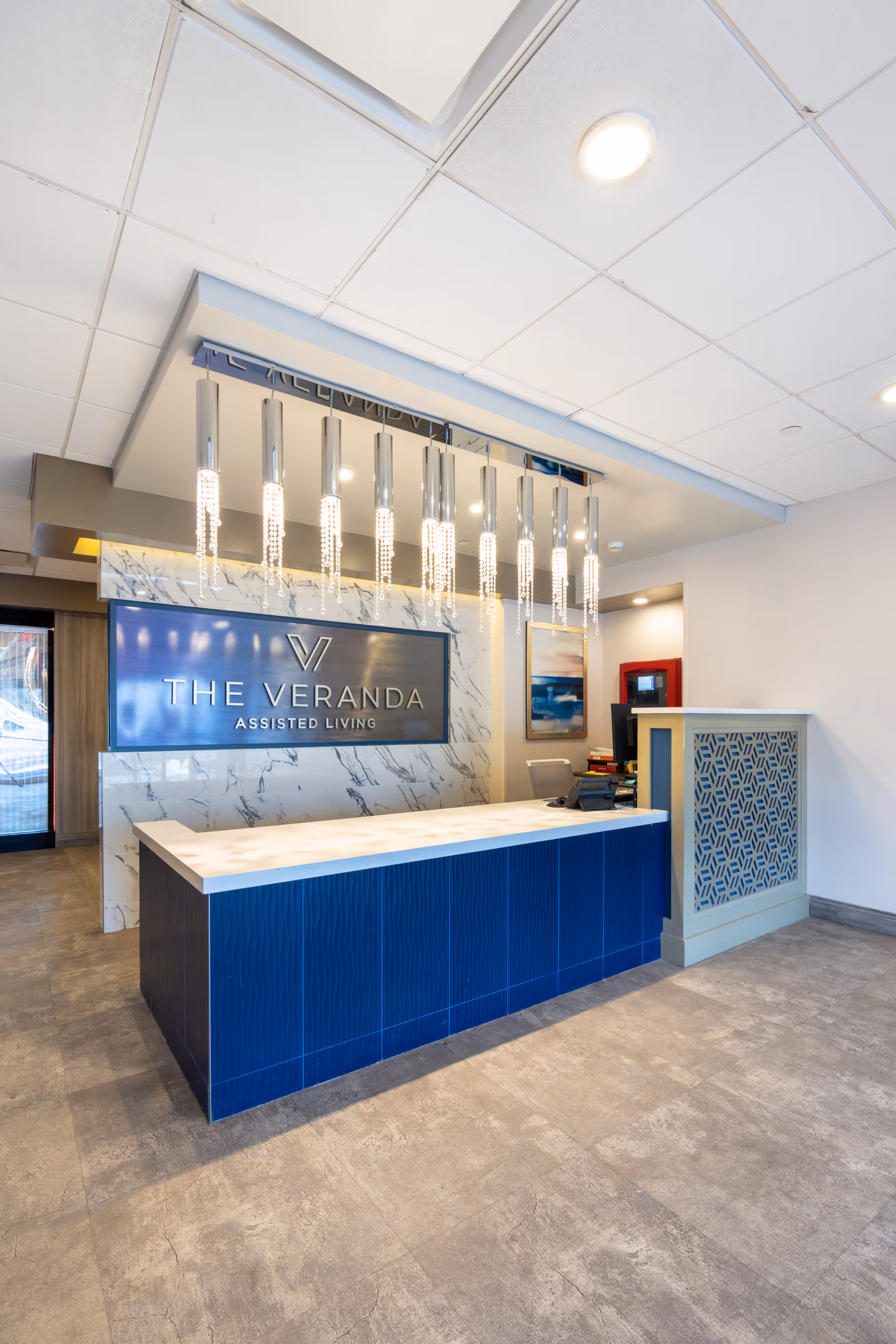Reception area of The Veranda Assisted Living facility featuring a modern blue and white front desk, a marble-patterned wall with the facility's name and logo, and a contemporary chandelier with hanging lights.