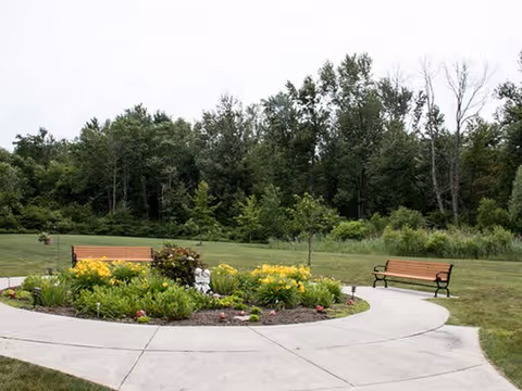 A circular concrete pathway surrounding a flower bed with yellow flowers and green plants, flanked by two wooden benches with black metal armrests and legs. In the background, there is a grassy area and a dense line of trees under an overcast sky.