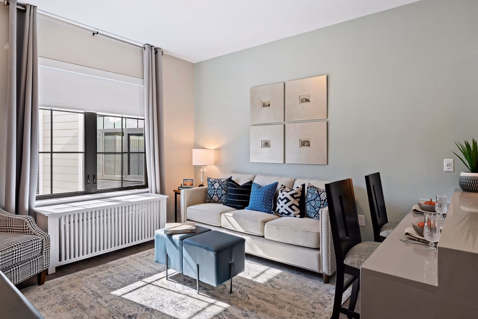 A bright and cozy living room with a beige sofa adorned with various patterned blue and white throw pillows. In front of the sofa are two small blue ottomans with a book on top. To the left, there is a large window with gray curtains and a white radiator cover beneath it. A patterned armchair is partially visible on the left side. On the right, a kitchen counter with two black chairs is set with plates, bowls, and glasses. The walls are painted light gray with four framed pictures hanging above the sofa. A table lamp is on a small side table next to the sofa.