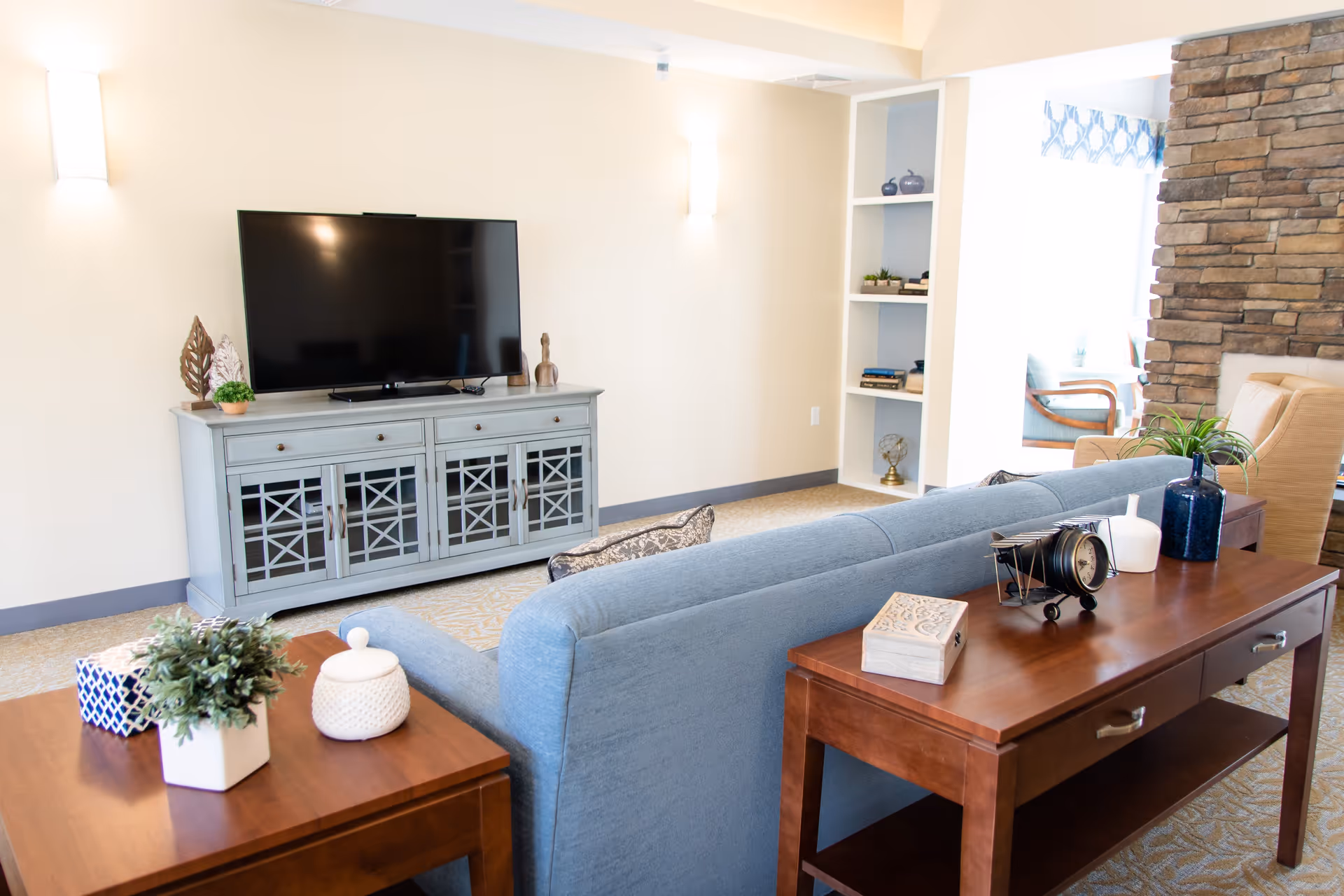 Bright seating area with a blue couch facing a flat-screen TV on a gray media console, wooden side tables, and decorative accents.