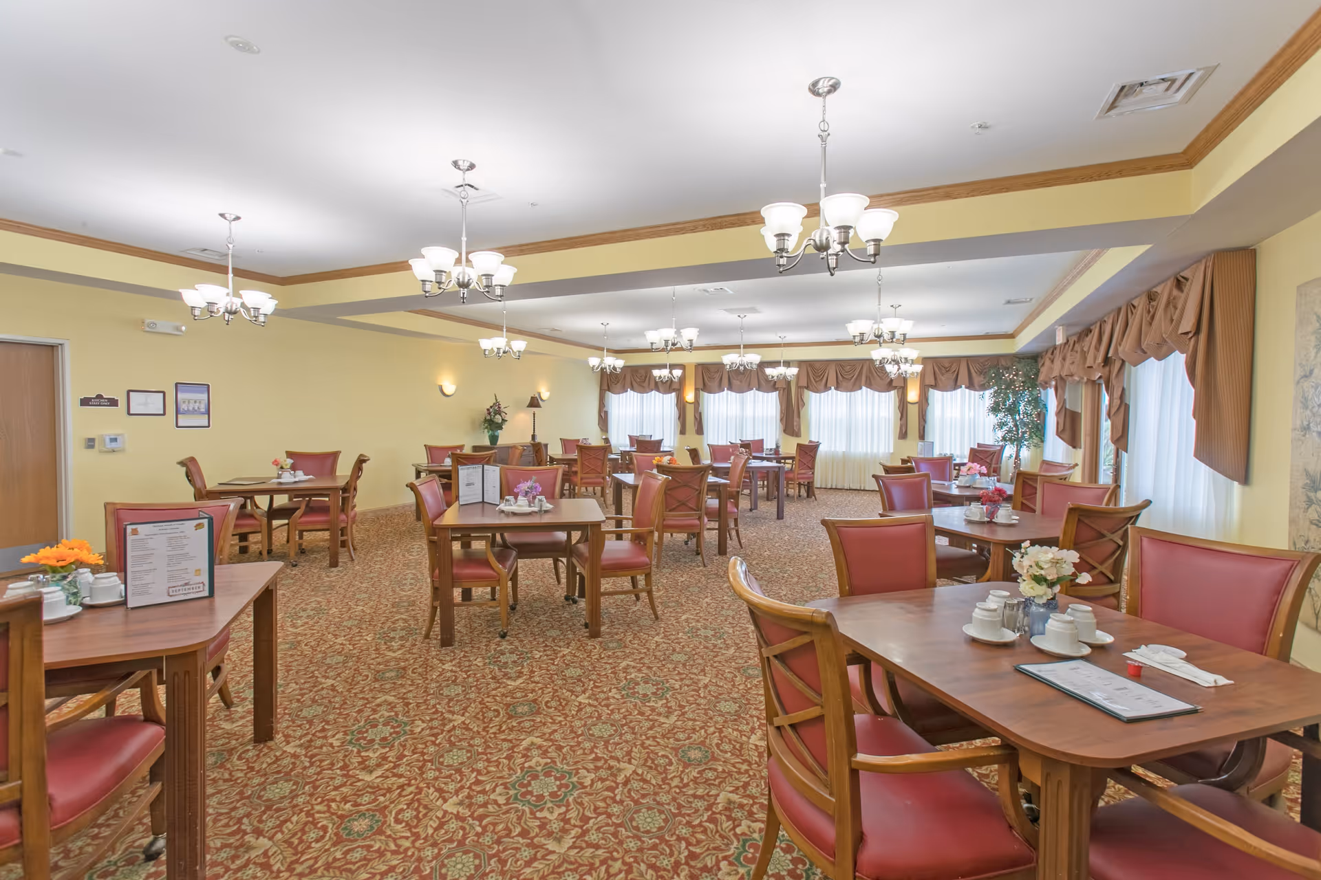 A spacious dining room with multiple wooden tables and red cushioned chairs arranged neatly. The room has yellow walls, patterned carpet flooring, and several chandeliers hanging from the ceiling. Windows with curtains allow natural light to brighten the space, and there are small floral centerpieces on the tables.