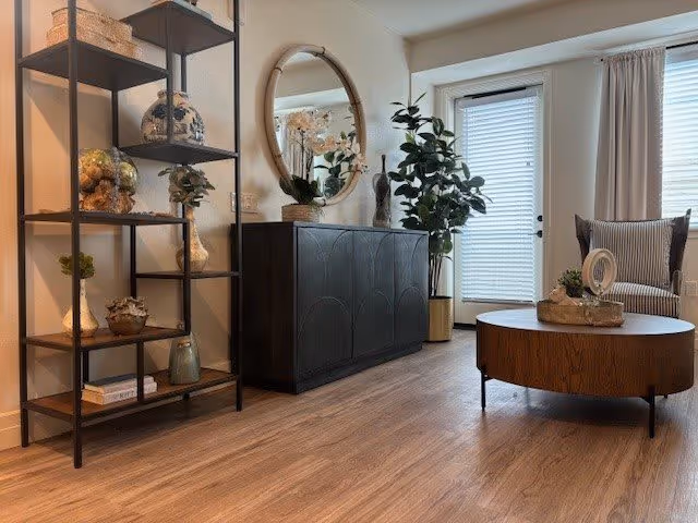 Stylish living room with shelving, a dark sideboard topped by a round mirror, potted plants, a round coffee table and an armchair by a glass door with blinds.