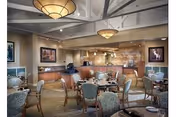 A spacious dining room with multiple round tables and cushioned chairs arranged neatly. The room features a high ceiling with exposed beams and two large hanging light fixtures. The back wall has a kitchen or serving area with wooden cabinetry and a countertop. Framed artwork is displayed on the walls, and the carpeted floor adds warmth to the space.