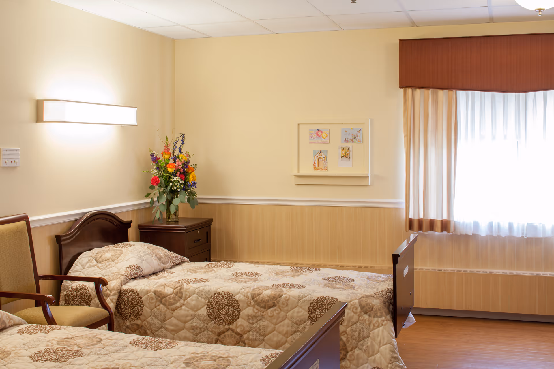 Two twin beds in a softly lit senior living bedroom with patterned bedspreads, a nightstand with a vase of flowers, a chair, and a curtained window.
