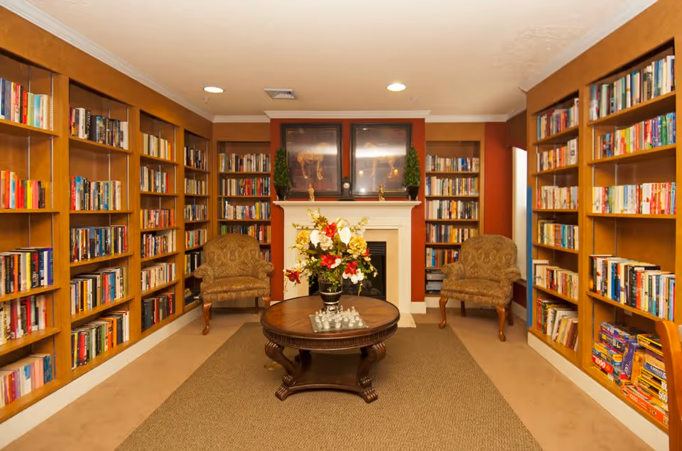 A cozy library room with wooden bookshelves filled with books lining both side walls. Two upholstered armchairs are placed near a white fireplace with a mantel decorated with two small potted plants and framed artwork above. A round wooden coffee table with a floral arrangement and a chess set sits on a beige carpet in the center of the room.