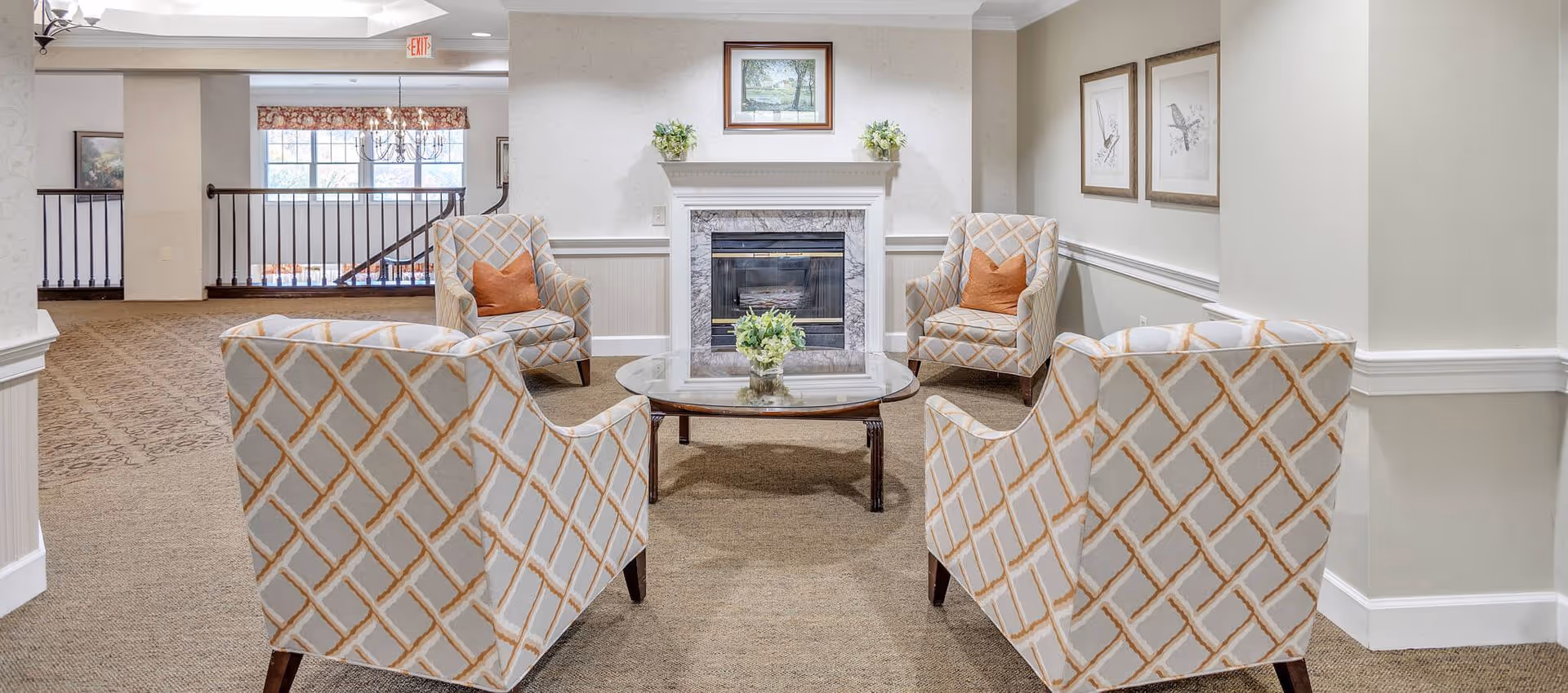 A cozy sitting area in a senior living facility with four patterned armchairs arranged around a glass coffee table. Behind the chairs is a white fireplace with a marble surround, decorated with two small plants and a framed picture above it. The walls are light-colored with white wainscoting, and there are two framed bird illustrations on the right wall. In the background, there is a railing and a window with a floral valance, allowing natural light into the space.