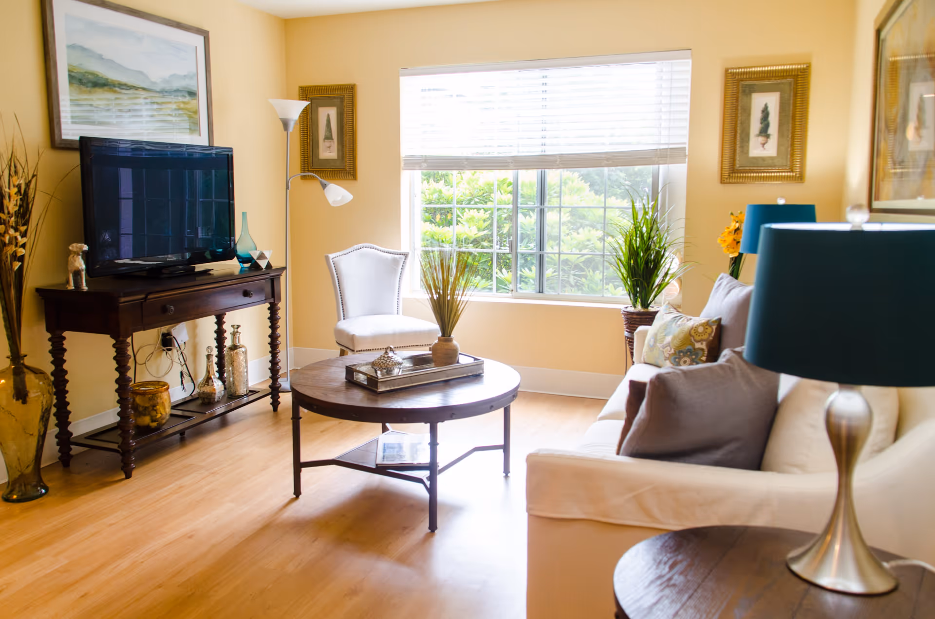A cozy living room with a large window letting in natural light. The room features a white sofa with decorative pillows, a round wooden coffee table with a tray and vase, a white upholstered chair, a wooden TV stand with a flat-screen TV, and various decorative items including framed artwork, plants, and lamps with teal shades.