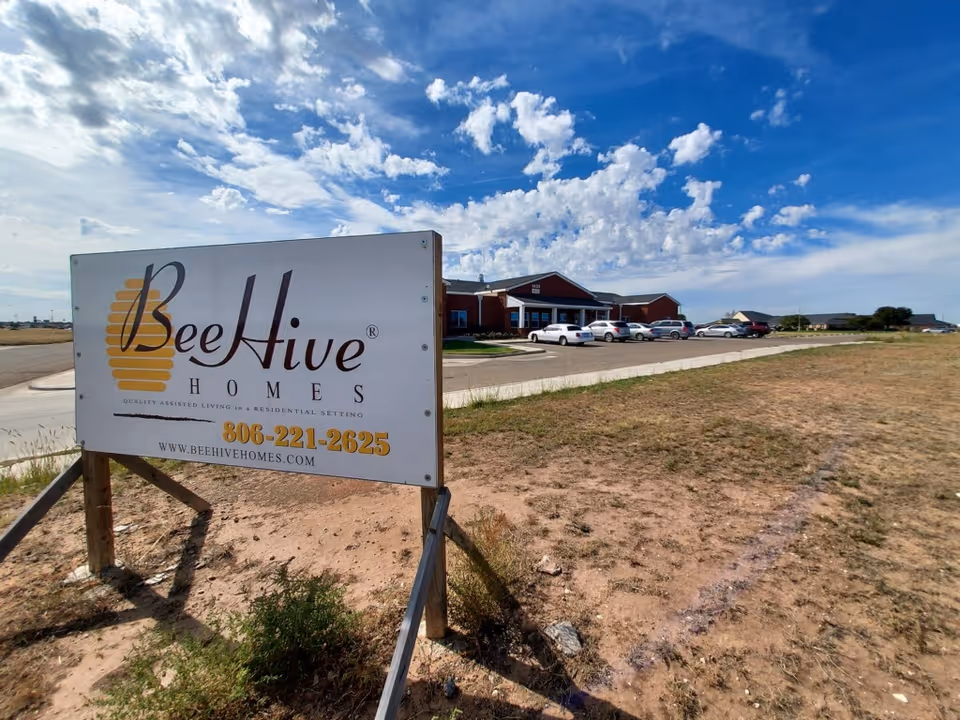 Outdoor view of BeeHive Homes of Plainview facility with a large sign in the foreground displaying the facility name, phone number, and website. The building and parking lot with several cars are visible in the background under a partly cloudy blue sky.