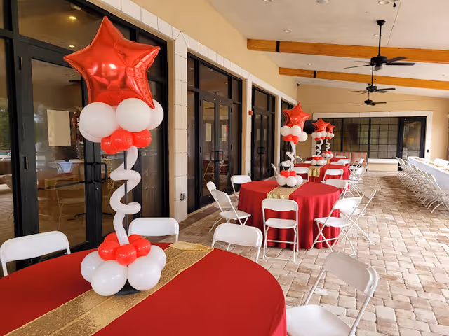Covered outdoor patio area with round tables covered in red tablecloths and gold runners. Each table has a centerpiece made of red and white balloons topped with a large red star-shaped balloon. White folding chairs are arranged around the tables. Ceiling fans are mounted on the ceiling with exposed wooden beams.