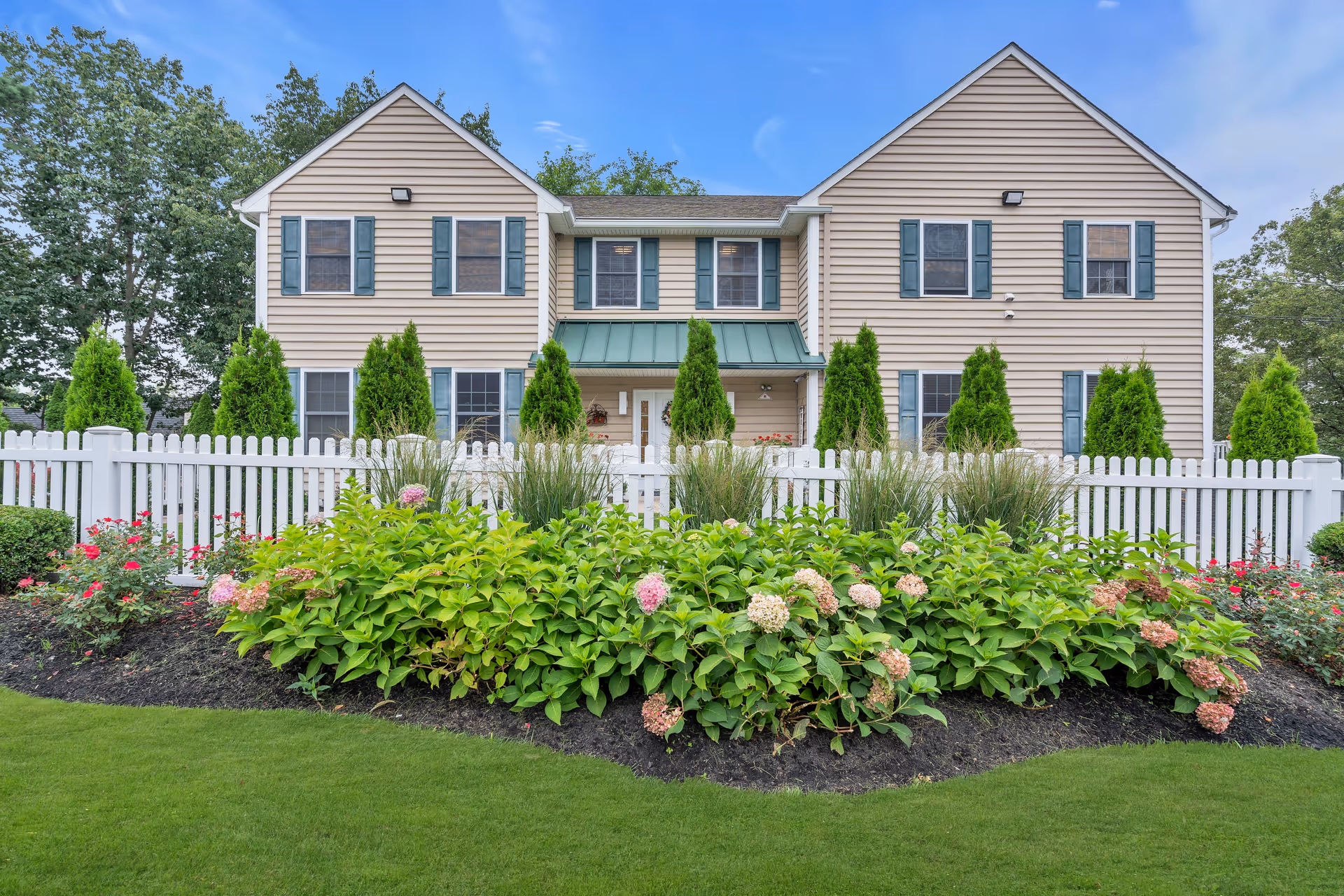 Two-story beige residential building with green shutters and a white picket fence, framed by flowering shrubs and a manicured lawn.