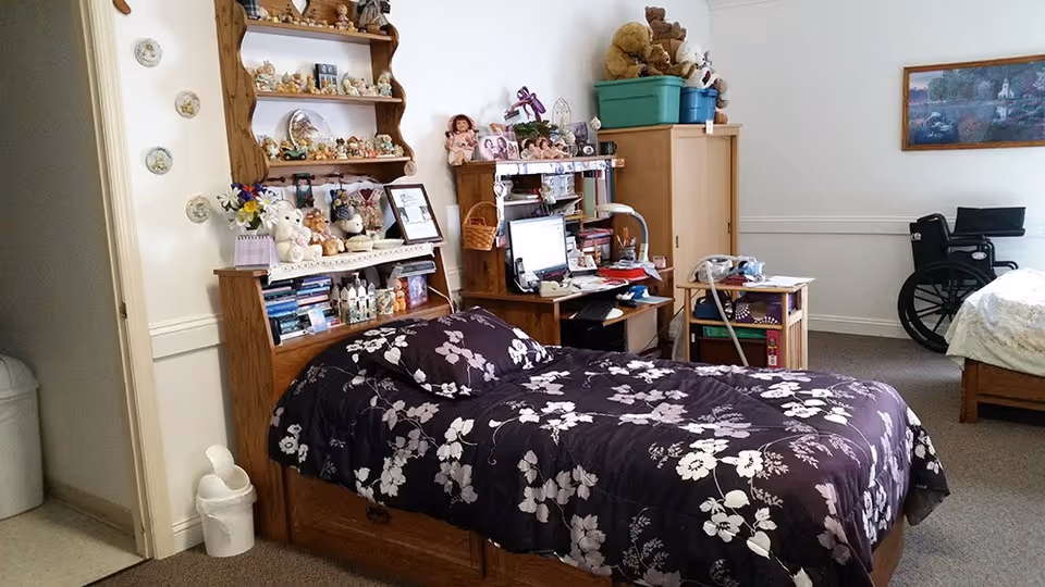 A furnished bedroom with a single bed covered in a dark floral comforter, shelving and a desk filled with personal items and stuffed animals, and a wheelchair near a second bed.