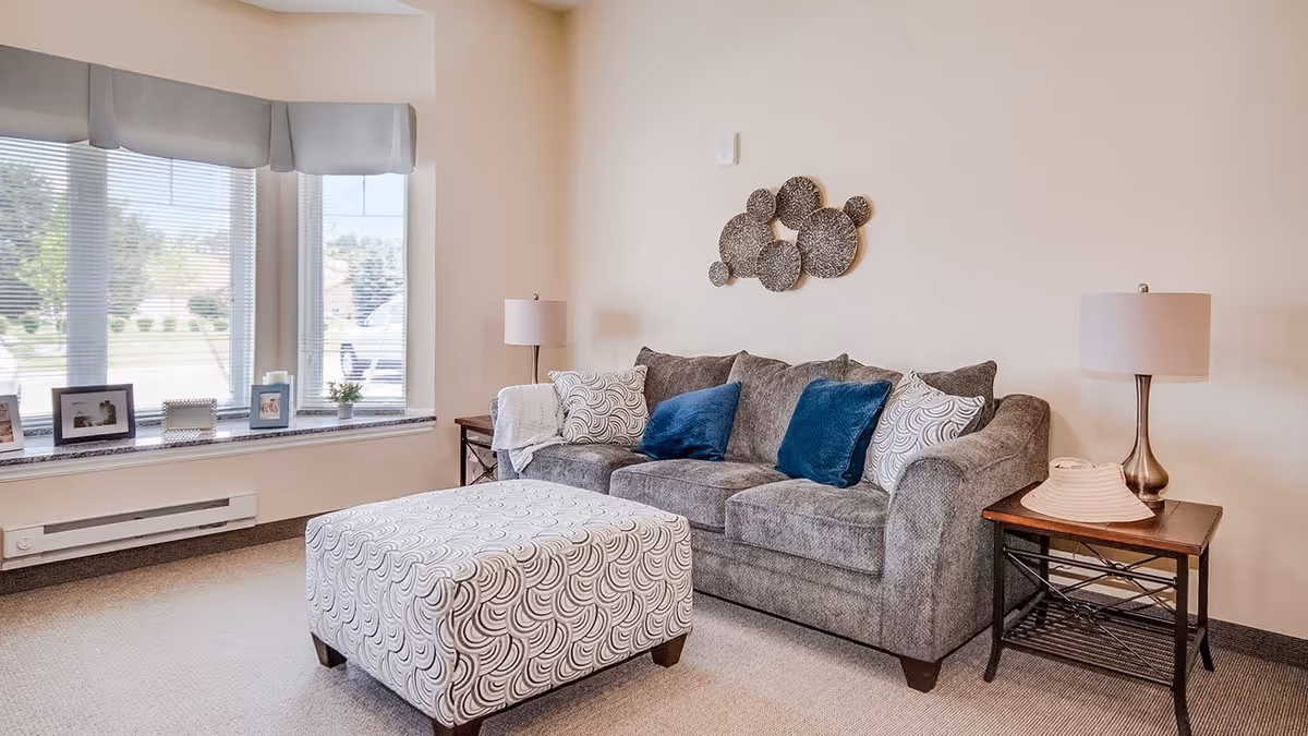 A cozy living room area with a gray sofa adorned with two blue and two patterned white pillows. In front of the sofa is a large patterned ottoman. On either side of the sofa are wooden side tables with lamps. A decorative wall art piece made of circular metal designs hangs above the sofa. A large window with blinds and a valance lets in natural light, with framed photos and small plants on the windowsill.