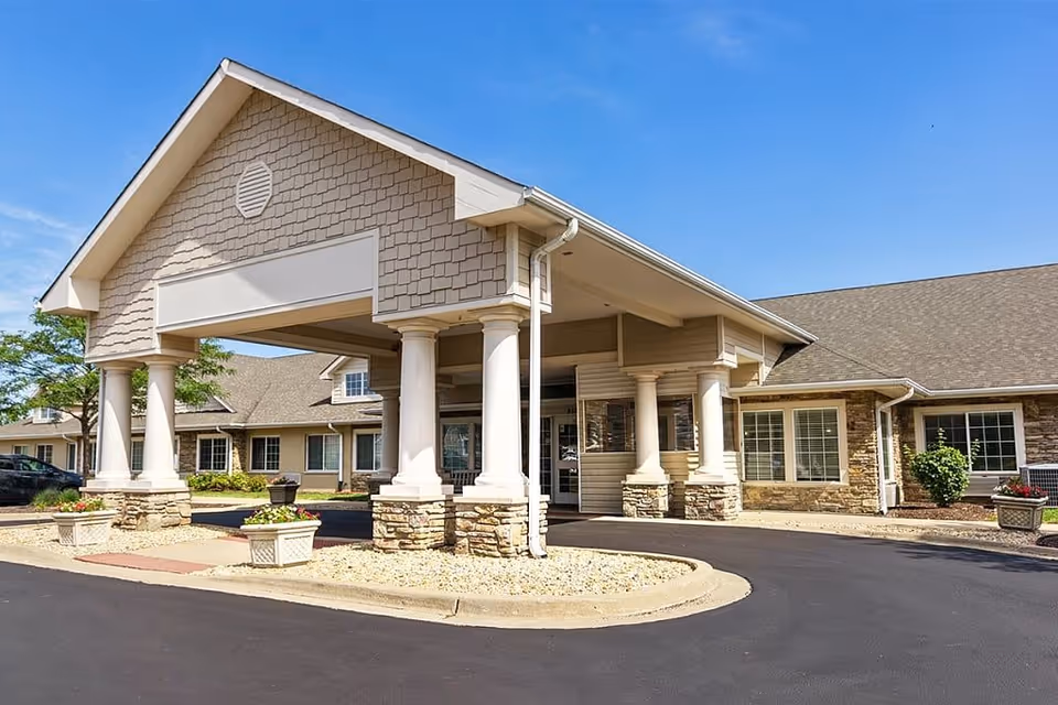 Covered entrance with white columns and planters at the front of a single-story senior living facility building under a blue sky.