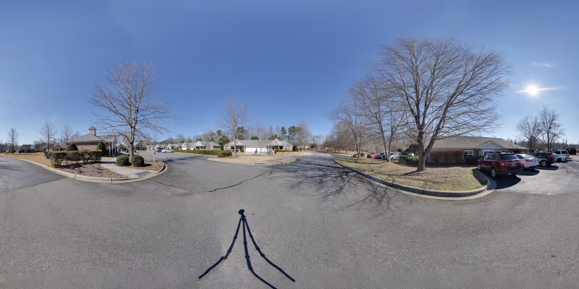 Wide panoramic view of the parking lot and surrounding buildings at Mint Hill Senior Living on a clear sunny day with leafless trees and several parked cars.