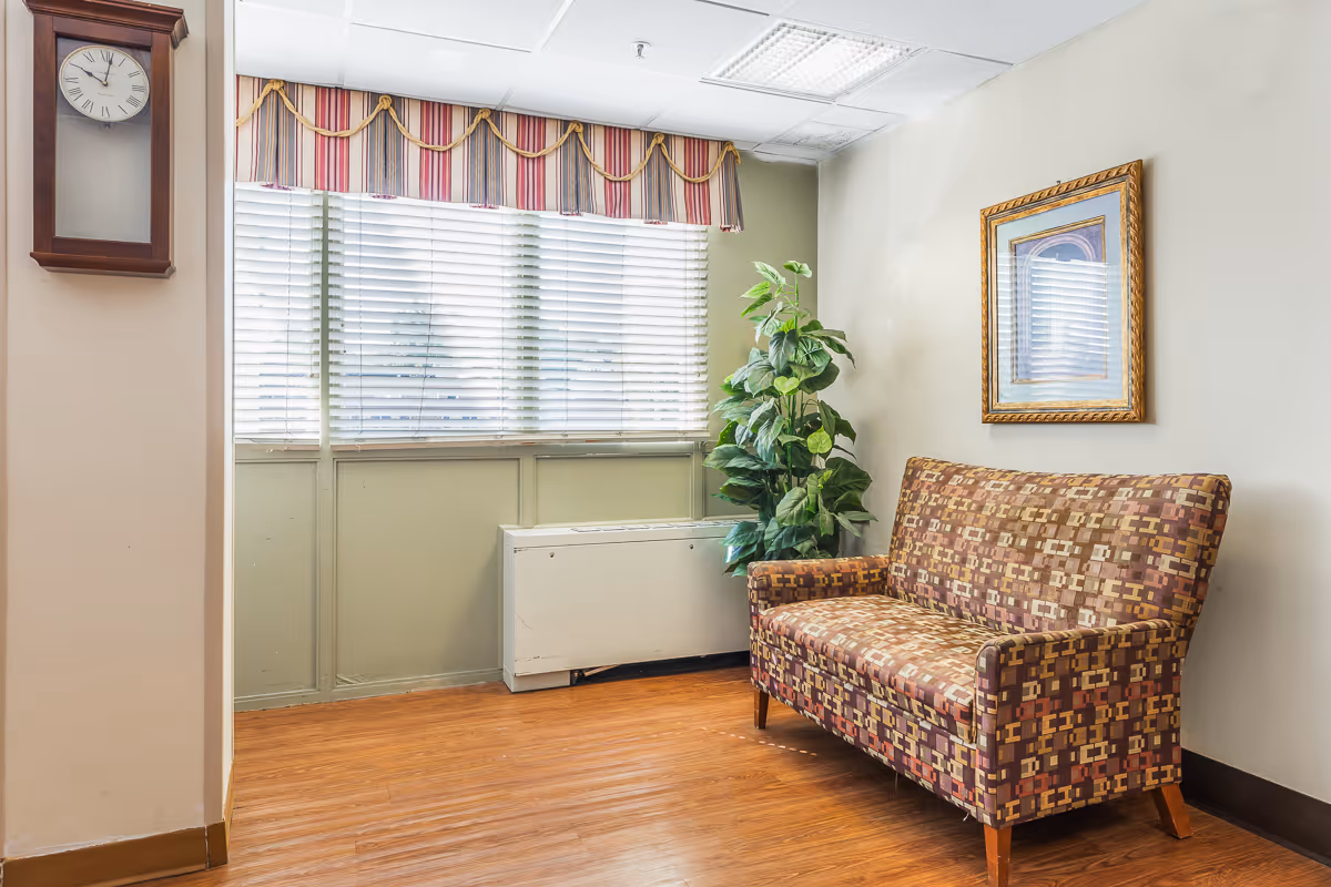 A small seating area with a patterned two-seater sofa, a potted plant, a framed picture on the wall, a wall clock, and a window with blinds and a striped valance. The floor is wooden and the walls are light-colored.