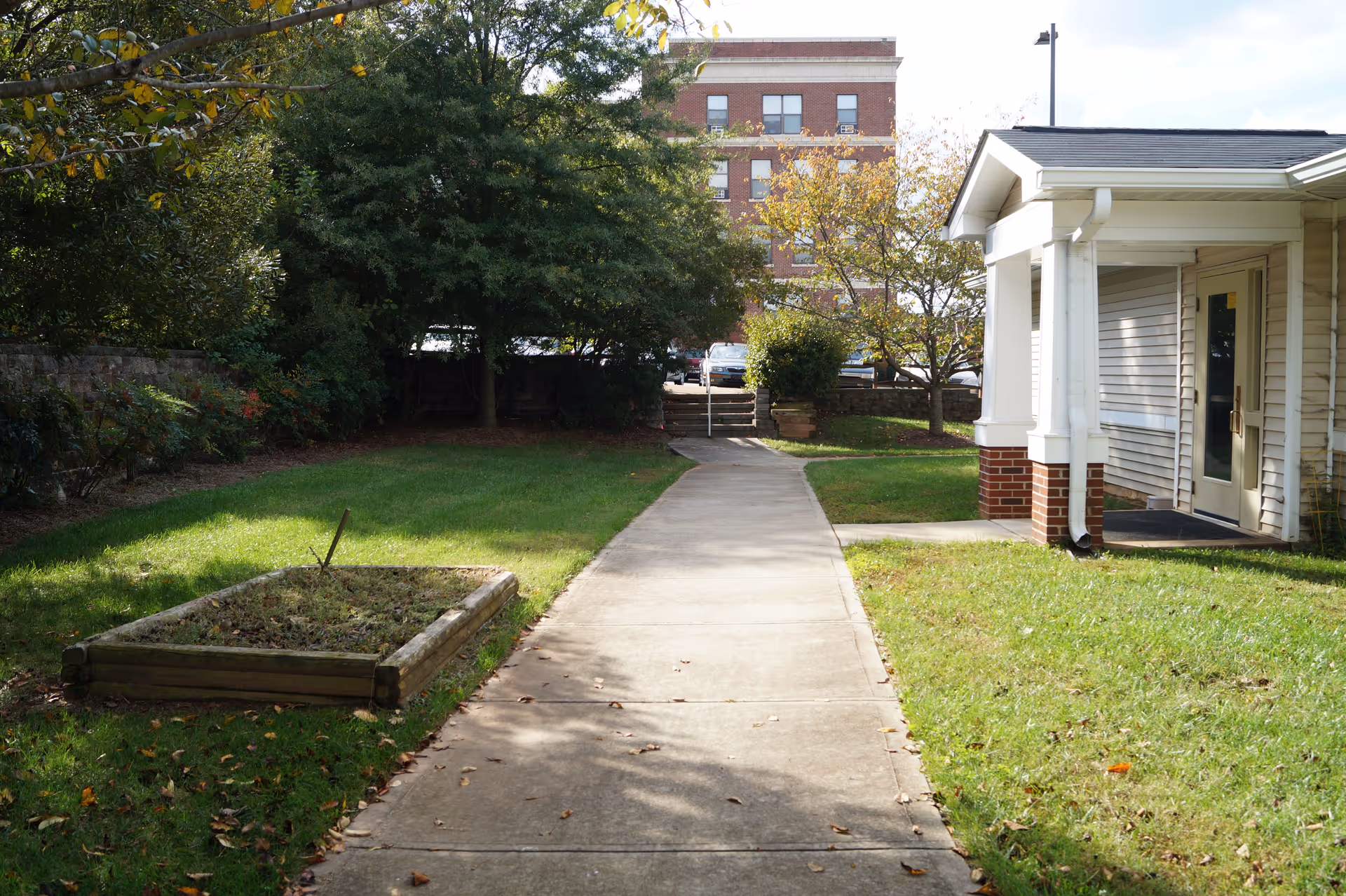 A paved walkway leading through a grassy outdoor area with trees and a small raised garden bed on the left. On the right is the corner of a building with white siding and brick columns. In the background, there is a multi-story brick building and some parked cars.