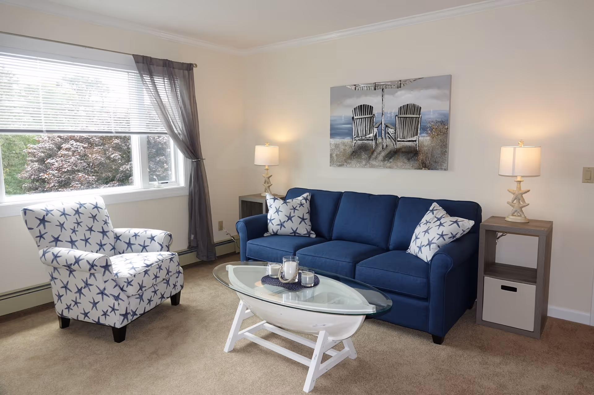 A cozy living room with a blue sofa adorned with starfish-patterned pillows, a matching armchair with a starfish pattern, a glass-top coffee table with candles, two side tables each with a lamp featuring a starfish base, and a large window with sheer curtains letting in natural light. A painting of two chairs facing the ocean hangs on the wall above the sofa.