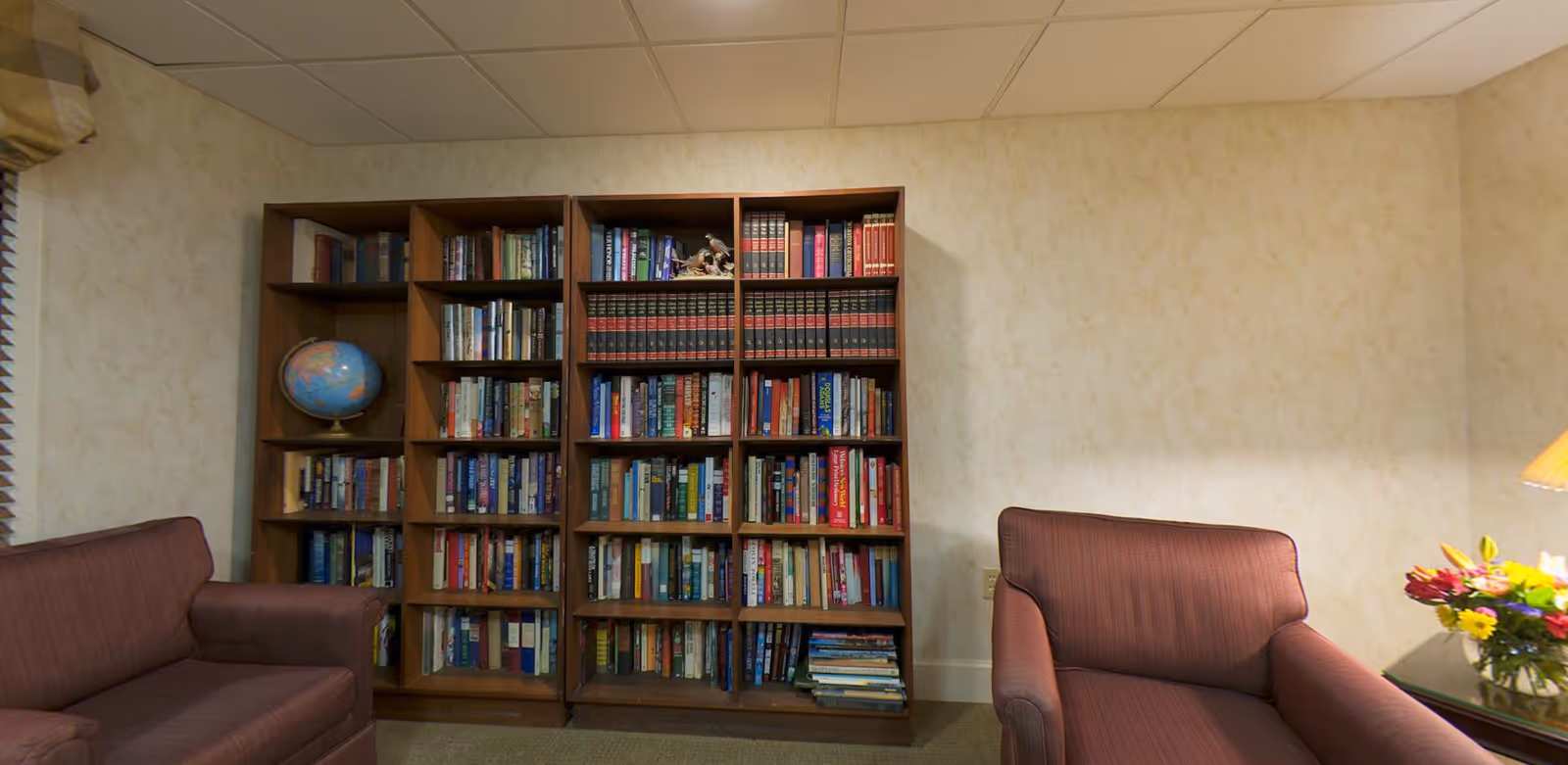 A cozy interior room with three wooden bookshelves filled with books and a globe on one shelf. There are two maroon upholstered armchairs on either side of the bookshelves. A table with a lamp and a colorful bouquet of flowers is visible on the right side. The walls are light-colored with a subtle pattern, and the ceiling has white tiles.