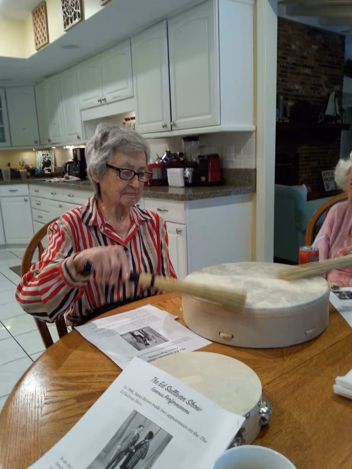 An elderly woman wearing glasses and a red and white striped shirt is sitting at a wooden table in a kitchen. She is playing a large drum with drumsticks. On the table, there are papers with text and images, a tambourine, and a cup. The kitchen has white cabinets and various appliances on the countertop. Another elderly person is partially visible sitting at the table.