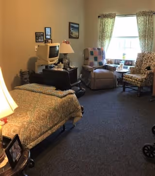 A cozy room with a single bed covered in a patterned bedspread, a small TV on a stand, a lamp, and a recliner chair with a colorful quilt draped over it. There is also a floral upholstered chair next to a window with patterned curtains, and a small table with decorative items including a small dog figurine.