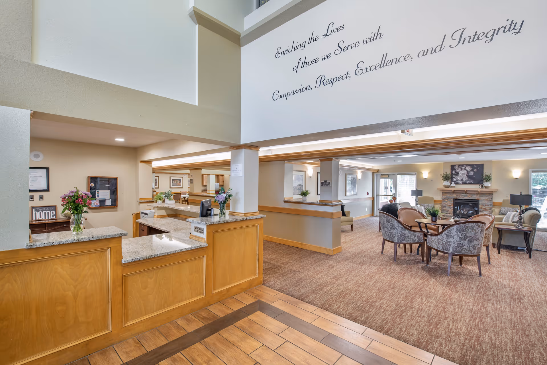 Interior view of a senior living facility reception area with a wooden front desk and granite countertop. Behind the desk, there are flowers and a small sign that says 'home'. The space opens into a comfortable living room area with upholstered chairs and sofas arranged around a coffee table and a stone fireplace. The walls are painted in neutral tones, and there is a large inspirational quote on the upper wall that reads 'Enriching the Lives of those we Serve with Compassion, Respect, Excellence, and Integrity'.