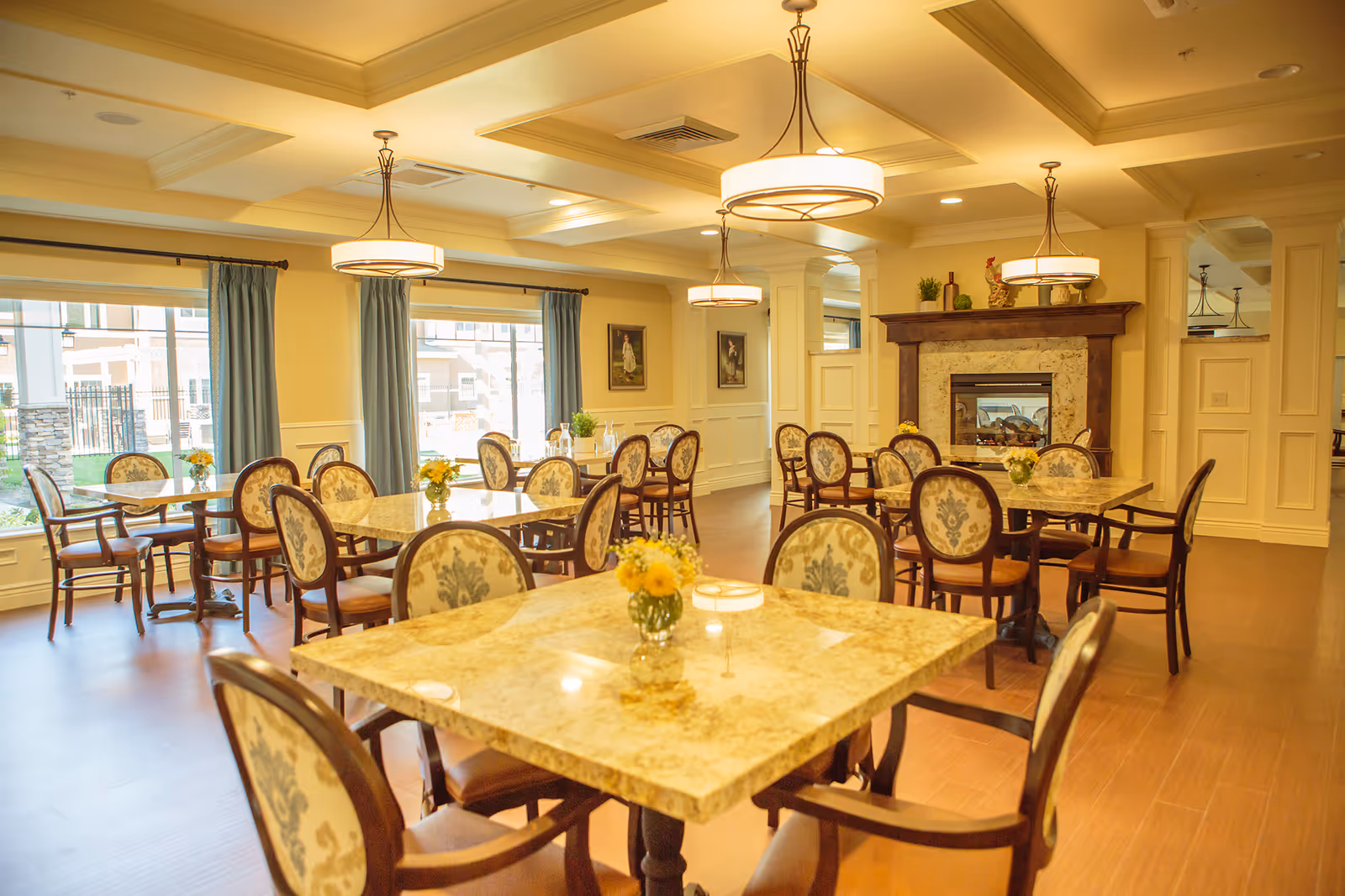 A bright and elegant dining room with multiple square marble tables surrounded by upholstered chairs with floral patterns. The room features large windows with blue curtains, a decorative fireplace with a wooden mantle, and warm lighting from ceiling fixtures. Small flower arrangements are placed on each table.