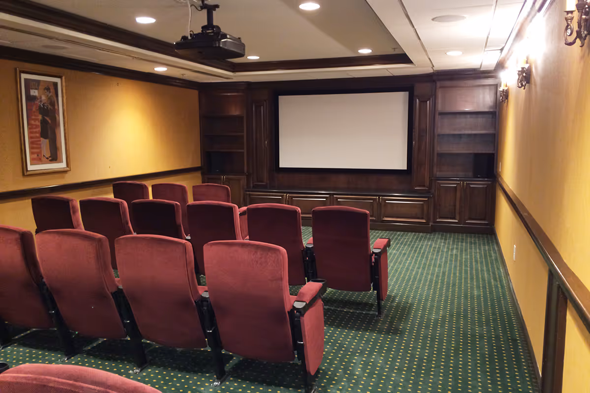 Small theater room with two rows of red upholstered seats facing a large blank projection screen. The room has yellow walls, green carpet with yellow dots, wooden built-in shelves and cabinets around the screen, and a ceiling-mounted projector.