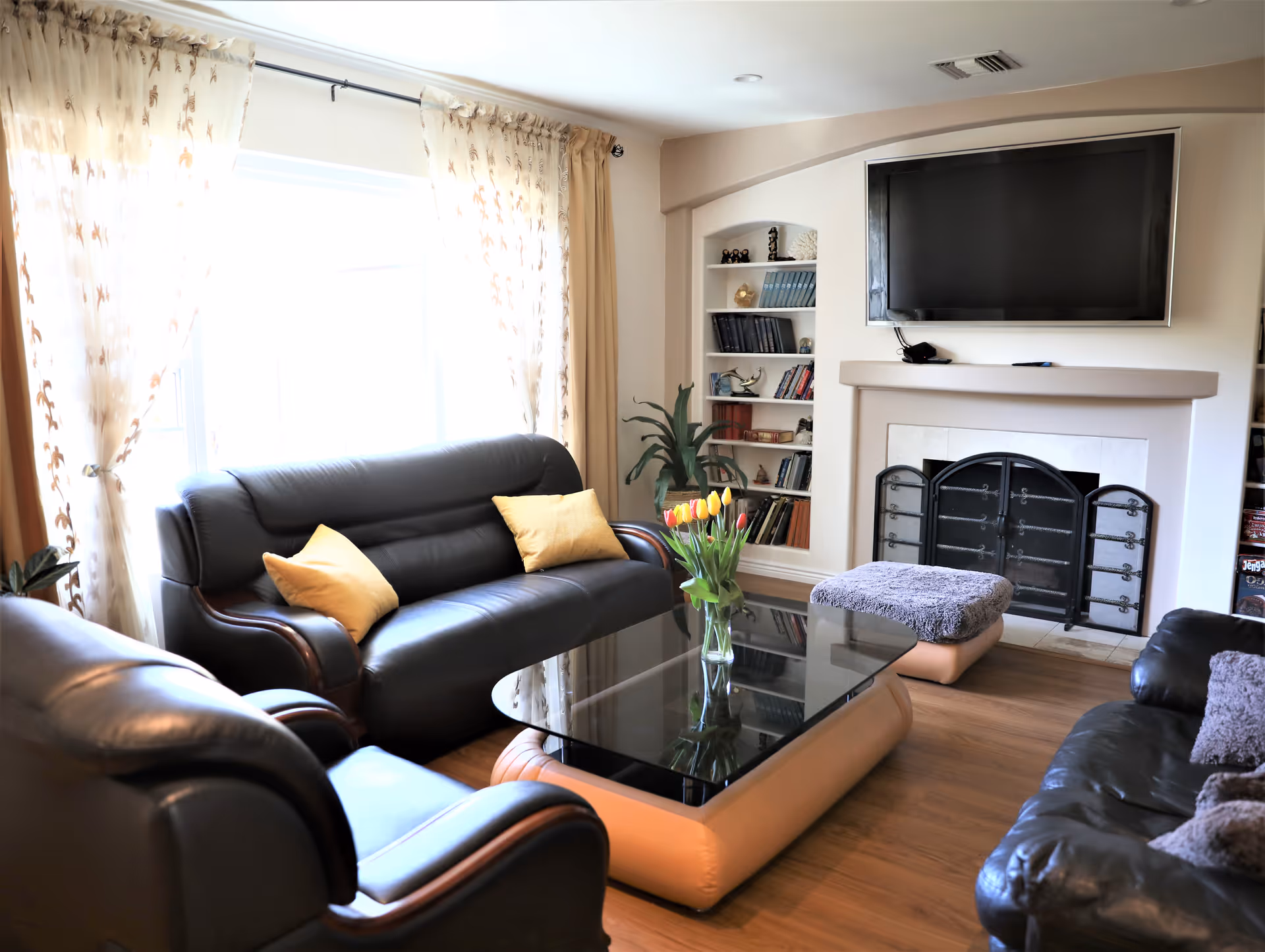 A bright living room with leather sofas around a glass coffee table holding a vase of tulips, a fireplace with a mounted TV, built-in bookshelves, and a large curtained window.