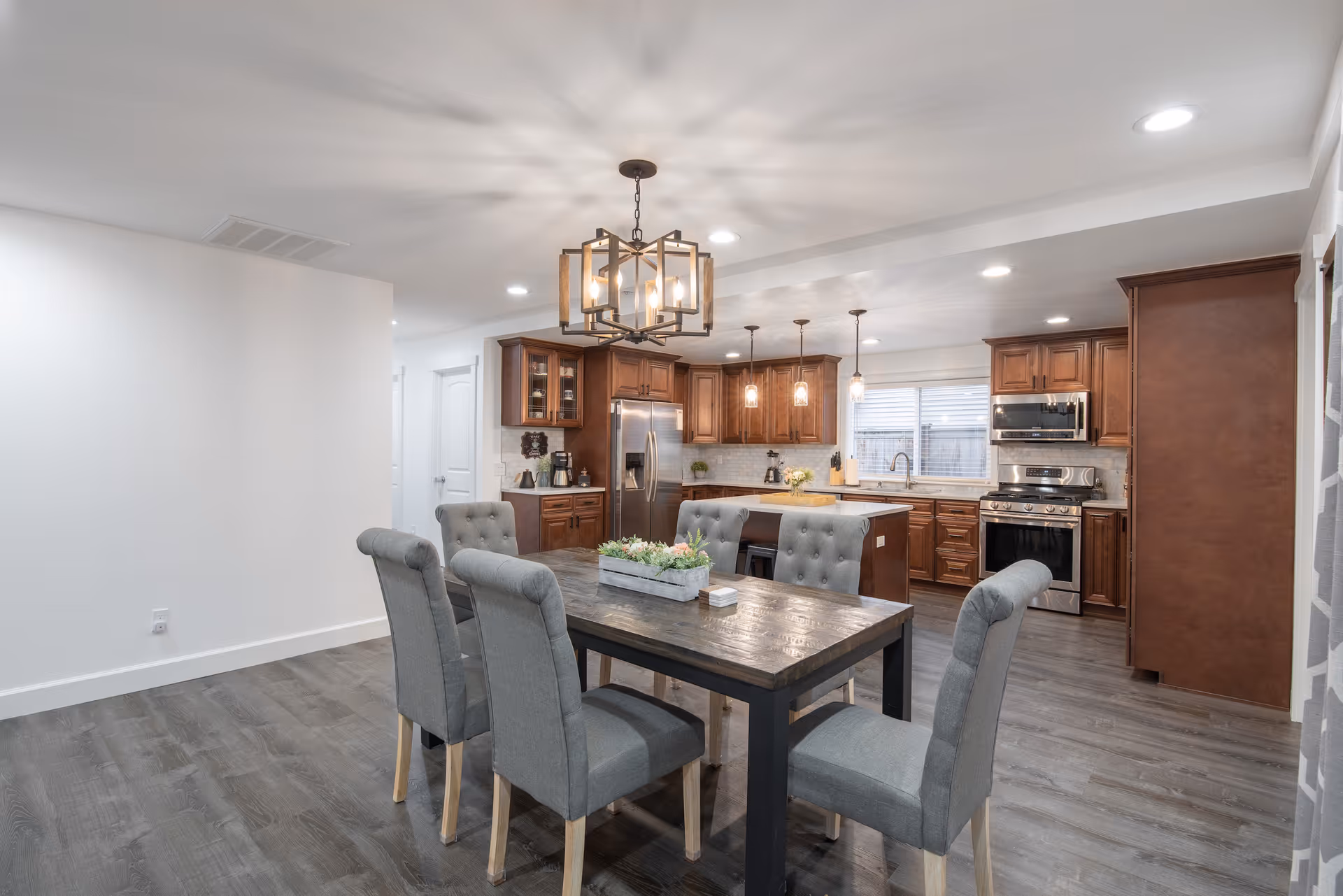 A modern kitchen and dining area with wooden cabinets, stainless steel appliances including a refrigerator, oven, and microwave. The dining table is dark wood with six gray upholstered chairs. Pendant lights hang above the kitchen island, and a decorative light fixture is above the dining table. The floor is a dark wood laminate, and the walls are painted white.