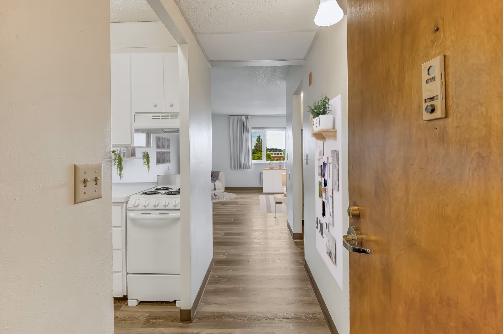 View from an open wooden door into a hallway of a senior living facility apartment. On the left, there is a small kitchen area with a white stove and cabinets. The hallway leads to a living room with a window showing greenery outside. The walls are light-colored, and there is a bulletin board with papers and a small plant on the right wall.