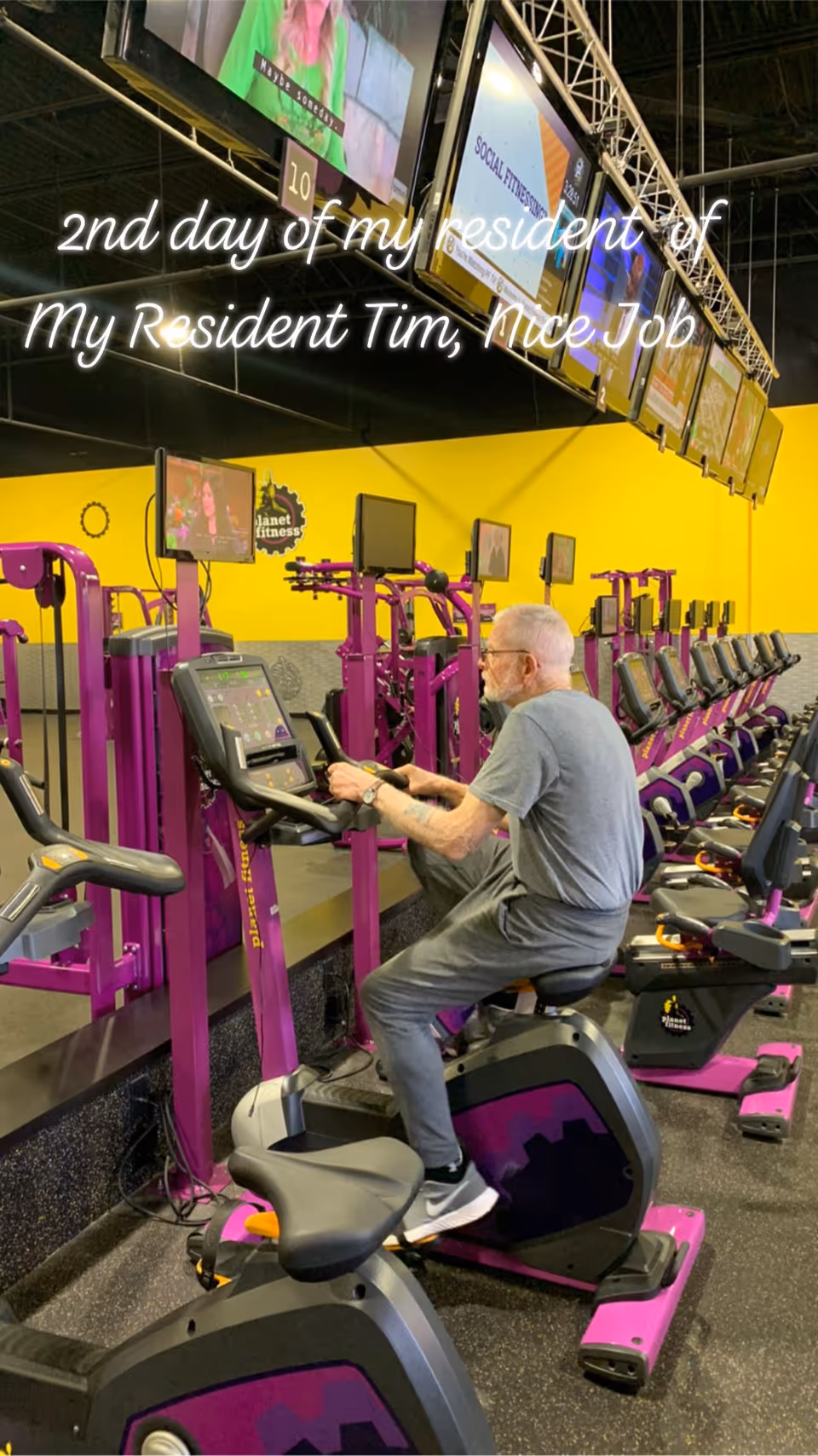 An elderly man exercising on a stationary bike in a gym with purple and black fitness equipment. Multiple screens are mounted above the equipment, displaying various content. The gym has bright yellow walls and a black ceiling.