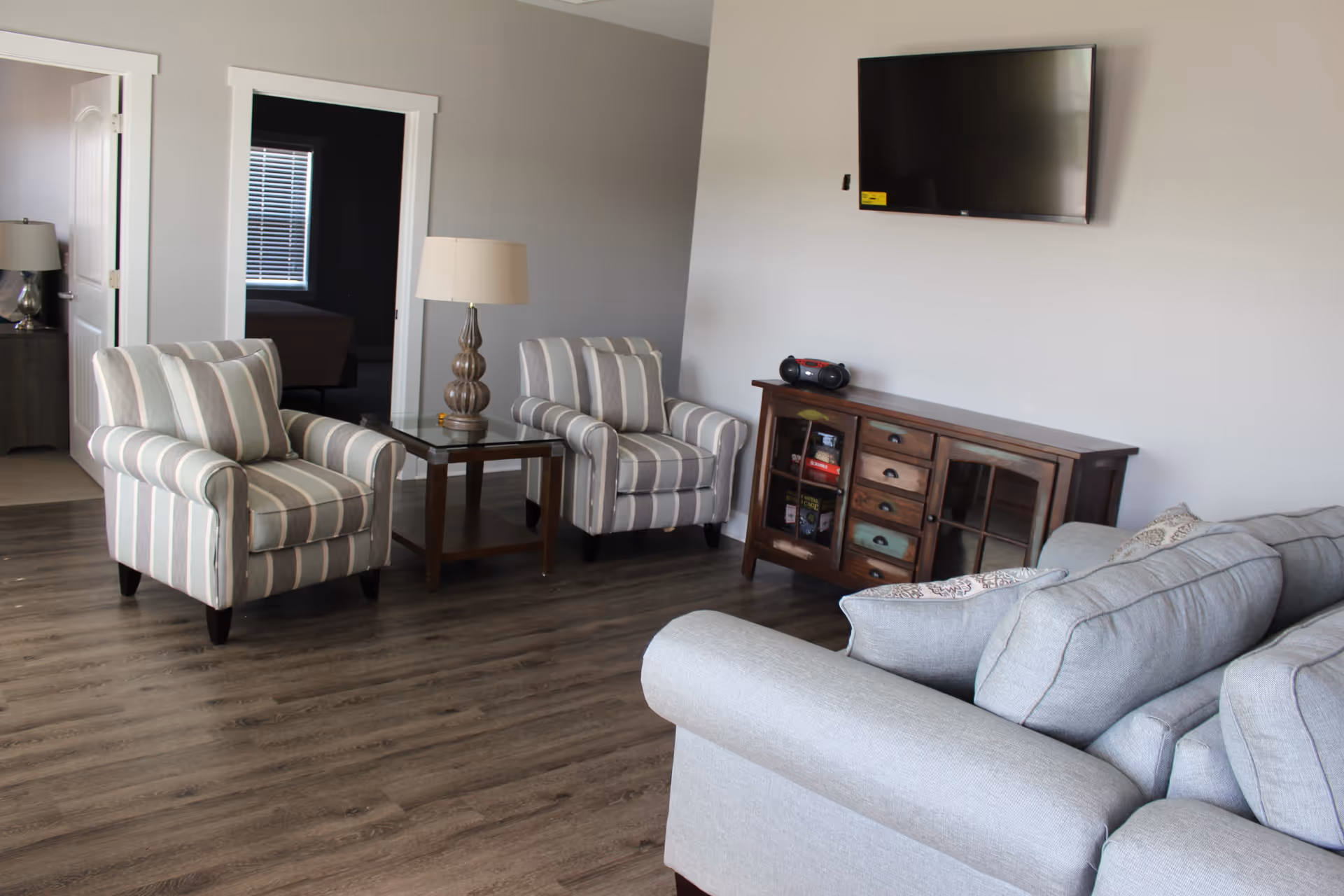 A living room with two striped armchairs, a glass-top side table with a lamp, a wooden cabinet with drawers and glass doors, a wall-mounted flat-screen TV, and a gray sofa. The room has wood flooring and neutral-colored walls.