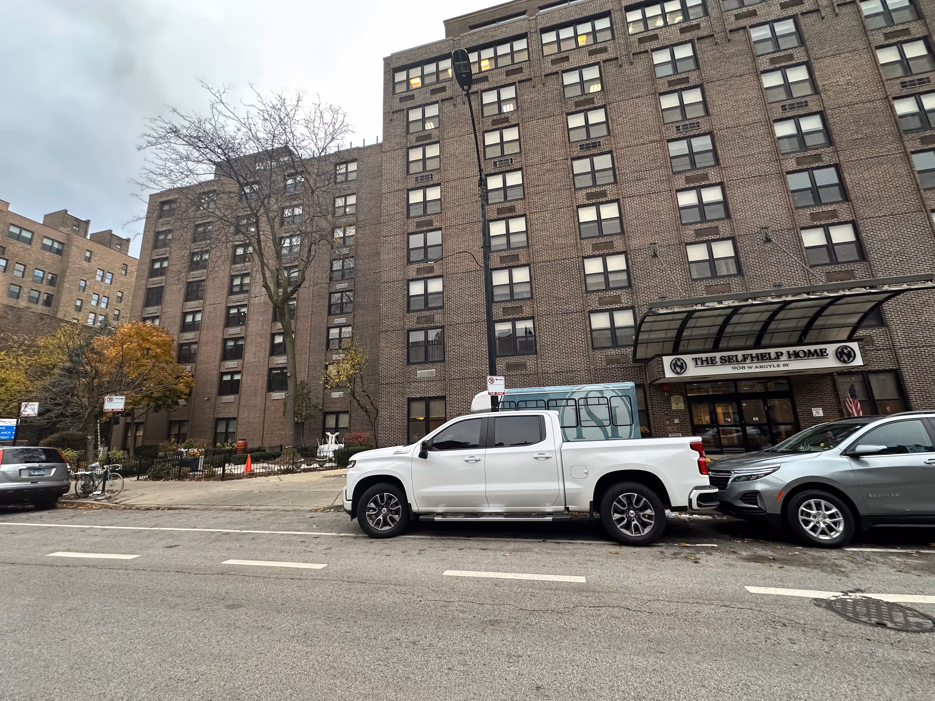 Street-level view of a multi-story brick senior living building with a canopy entrance reading "The Selfhelp Home" and cars parked in front.