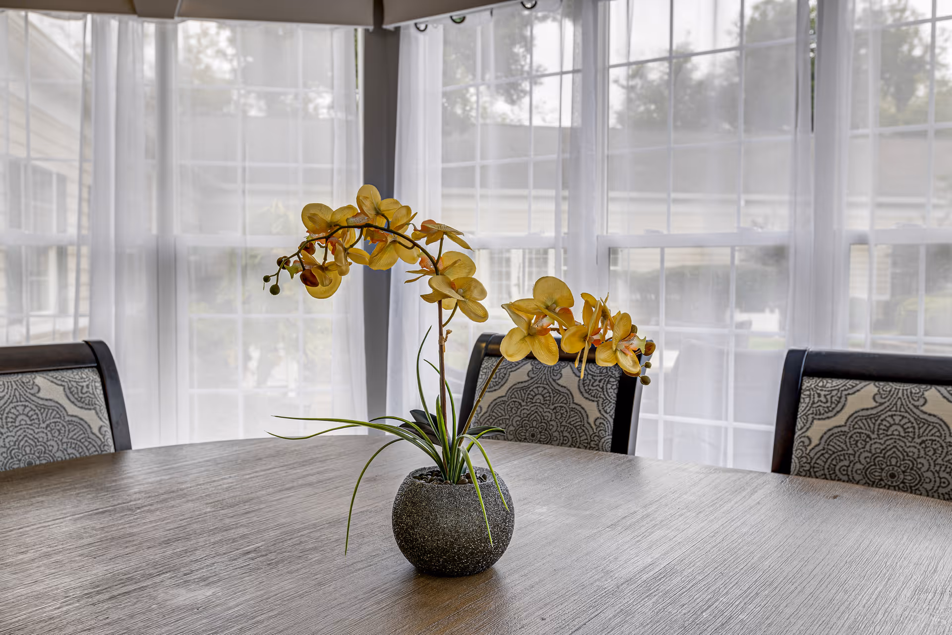 A round wooden dining table with a small gray pot holding yellow orchids in the center. Behind the table are chairs with patterned upholstery, and large windows with sheer white curtains letting in natural light.