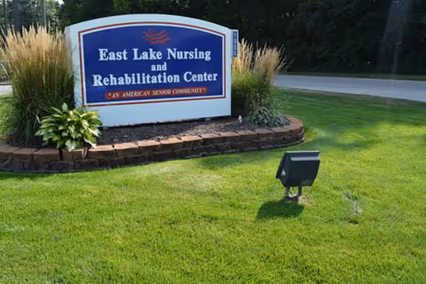 Entrance sign for East Lake Nursing and Rehabilitation Center on a landscaped lawn with ornamental grasses and a spotlight.