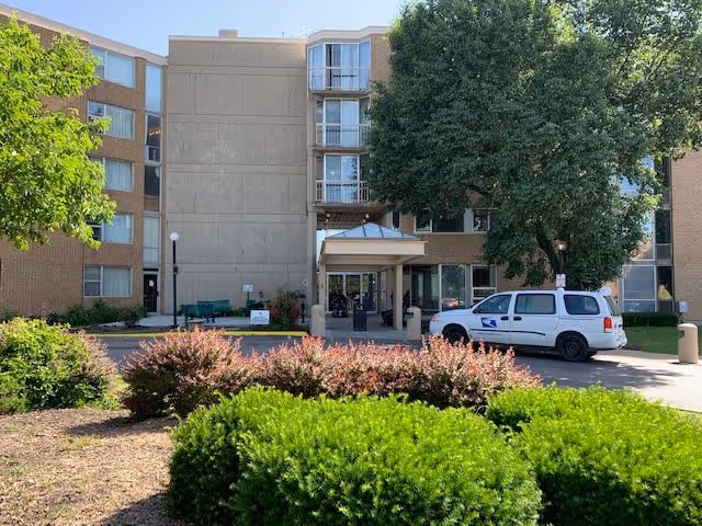 Exterior view of a multi-story residential building with a covered entrance. There are bushes and trees in the foreground, a white postal service vehicle parked near the entrance, and a clear blue sky above.