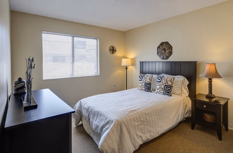 A neatly made bed with a black headboard and three decorative pillows in a bedroom. There is a black nightstand with a lamp on the right side of the bed and a floor lamp in the corner. A black dresser with decorative items is on the left side of the room, and a window with blinds is on the wall behind the bed.