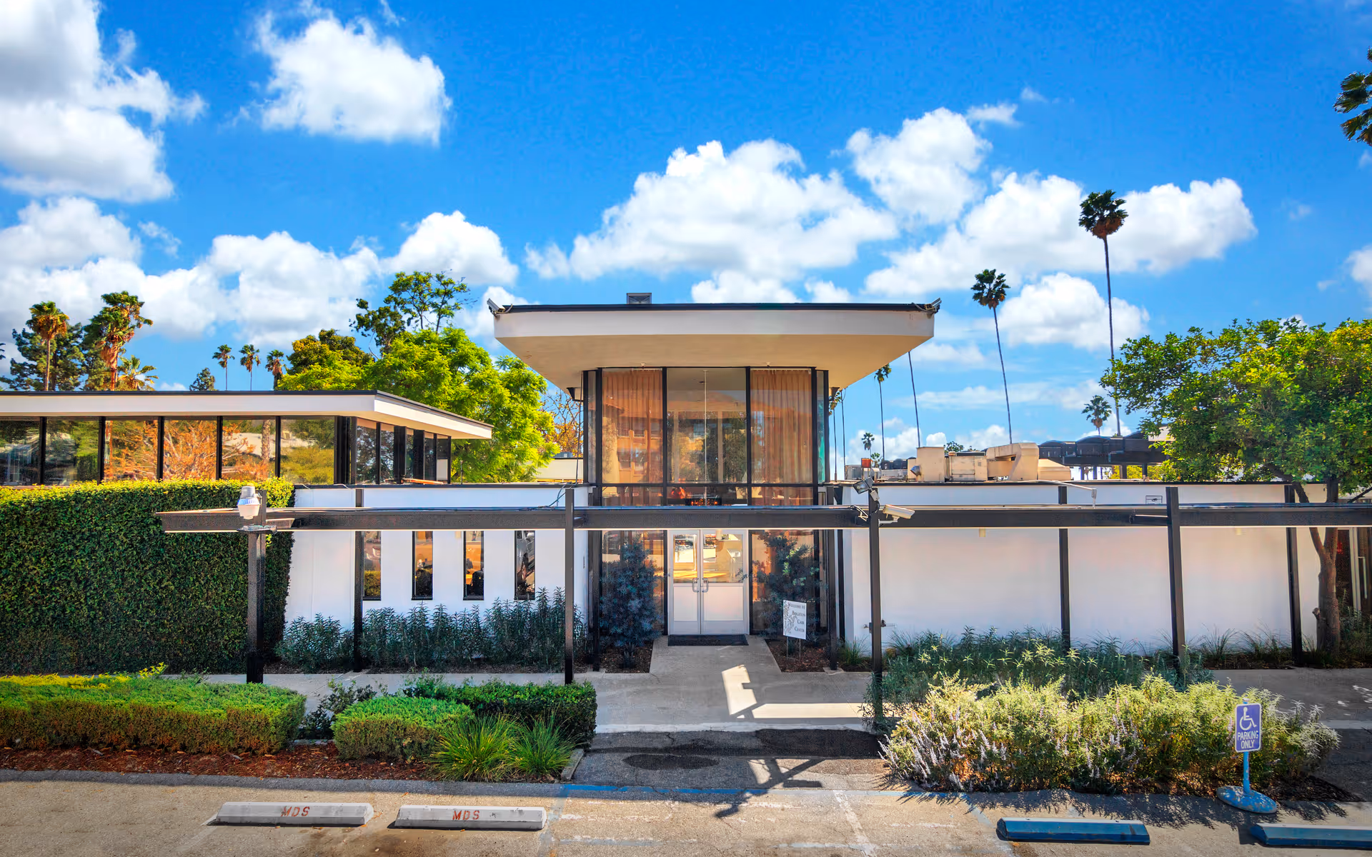 Front exterior of a modern low-rise building with a covered entrance, landscaped shrubs, and palm trees under a bright blue sky.