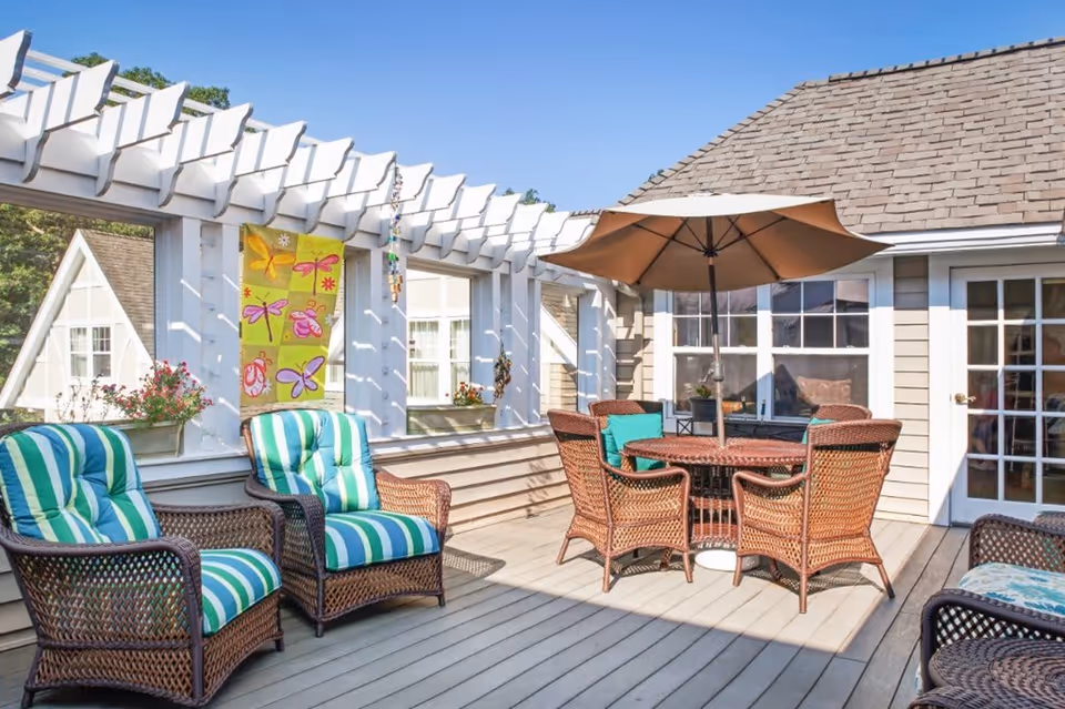 Outdoor patio area with wicker chairs featuring green and blue striped cushions, a round wicker table with an umbrella, and a decorative pergola with a colorful banner displaying dragonflies and flowers. The patio is attached to a building with beige siding and white-framed windows and doors.
