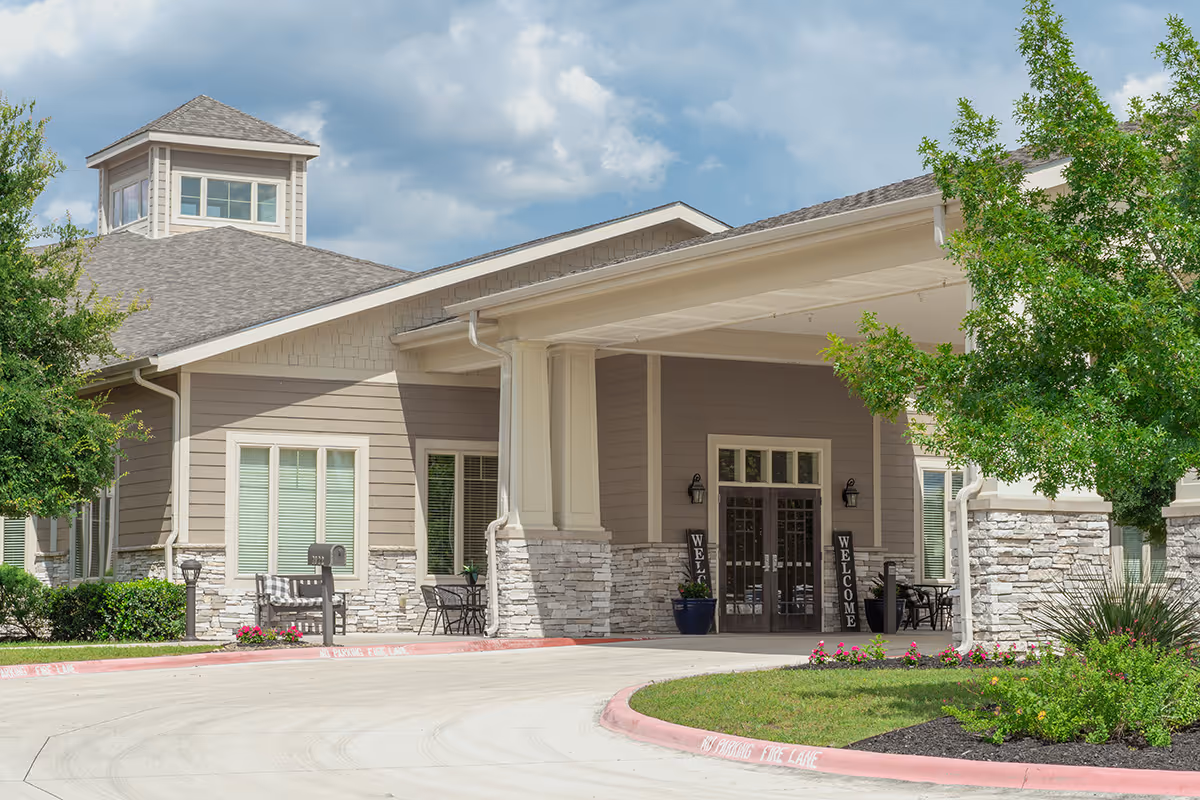 Front entrance of a senior living facility with a covered drop-off, stone accents, outdoor seating, and welcome signs.