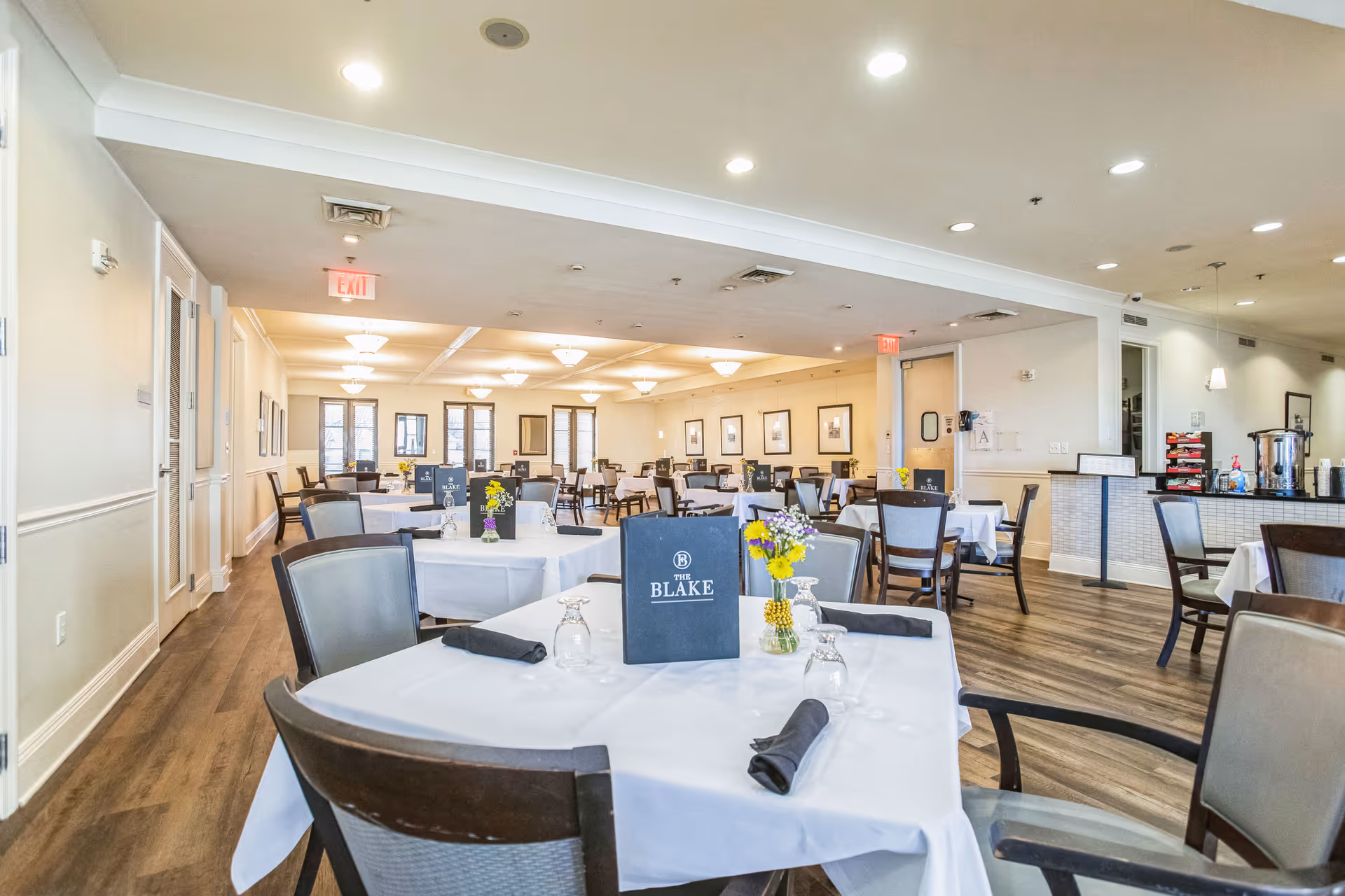 A spacious dining room with multiple tables covered in white tablecloths, each set with black napkins, upside-down glasses, and small flower arrangements. The room has wooden flooring, beige walls, and ceiling lights. Menus labeled 'The Blake' are placed on several tables.