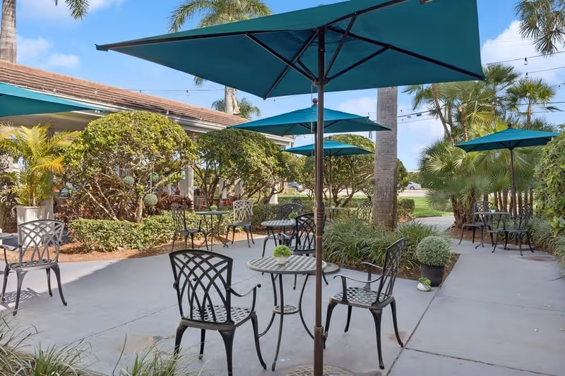 Outdoor patio area with several black metal tables and chairs under teal umbrellas, surrounded by trimmed bushes, palm trees, and other greenery on a sunny day.