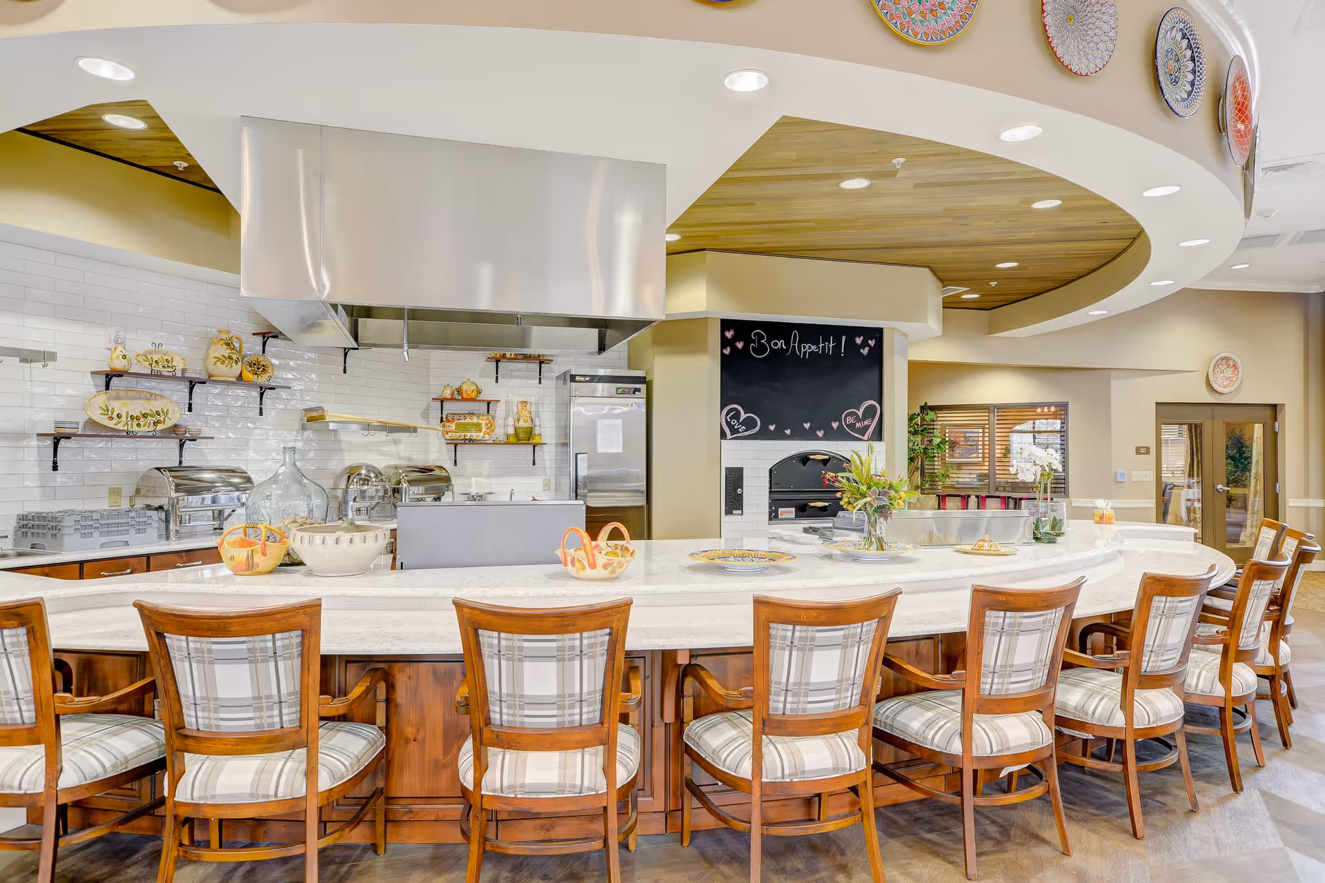 A bright and spacious kitchen area with a long curved marble countertop surrounded by wooden chairs with plaid cushions. The kitchen features stainless steel appliances, a large vent hood, and decorative shelves with ceramic dishes. A chalkboard on the wall reads 'Bon Appetit!' with heart decorations. The ceiling has recessed lighting and decorative plates are displayed along the curved wall.
