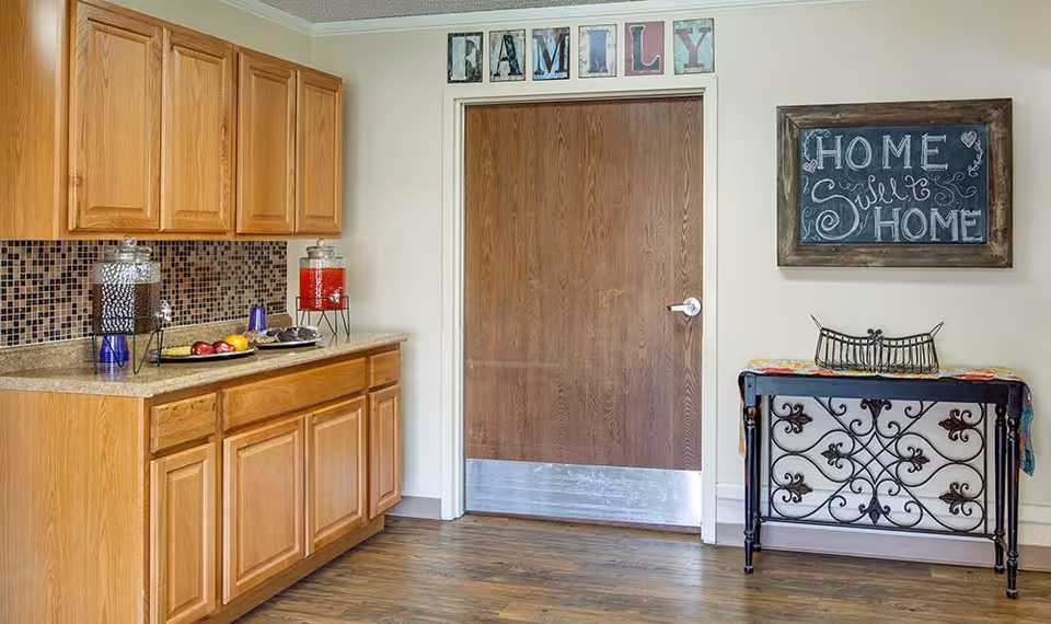 A small kitchenette/entry area with wooden cabinets and countertop drink dispensers, a central wooden door with a 'FAMILY' sign above, and a chalkboard reading 'Home Sweet Home' beside a decorative table.